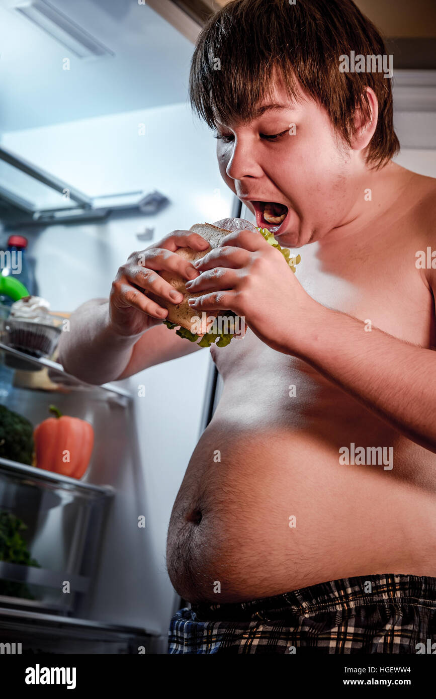 Faim fat man holding a big sandwich dans ses mains et se tenait à côté de l'Open d''un réfrigérateur. Aliments malsains. Banque D'Images