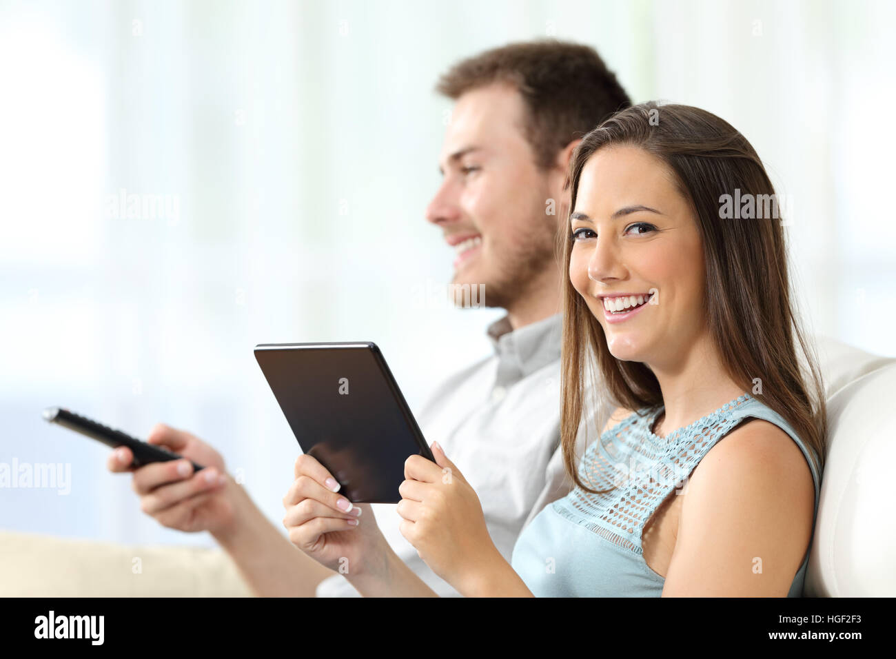 Couple watching tv assis sur un canapé dans le salon à la maison. L'homme est le zapping et woman is holding a tablet and looking at you Banque D'Images