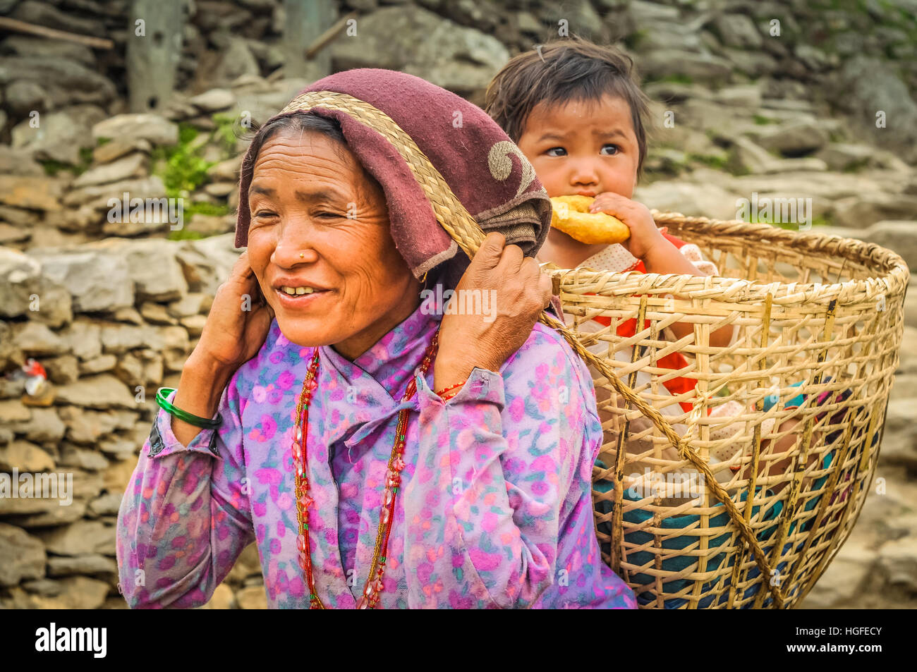 Beni, le Népal - circa Mai 2012 : femme autochtone de purple shirt porte panier sur son dos avec son enfant qui mange des gâteaux et elle marche jusqu'à la colline Banque D'Images