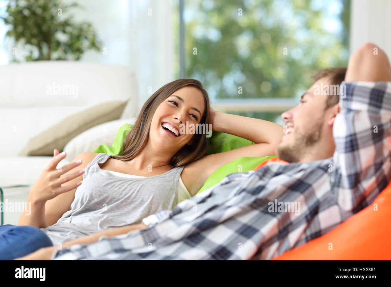 Couple ou colocataires parler et rire couché sur poufs dans la salle de séjour à la maison avec une fenêtre à l'arrière-plan Banque D'Images