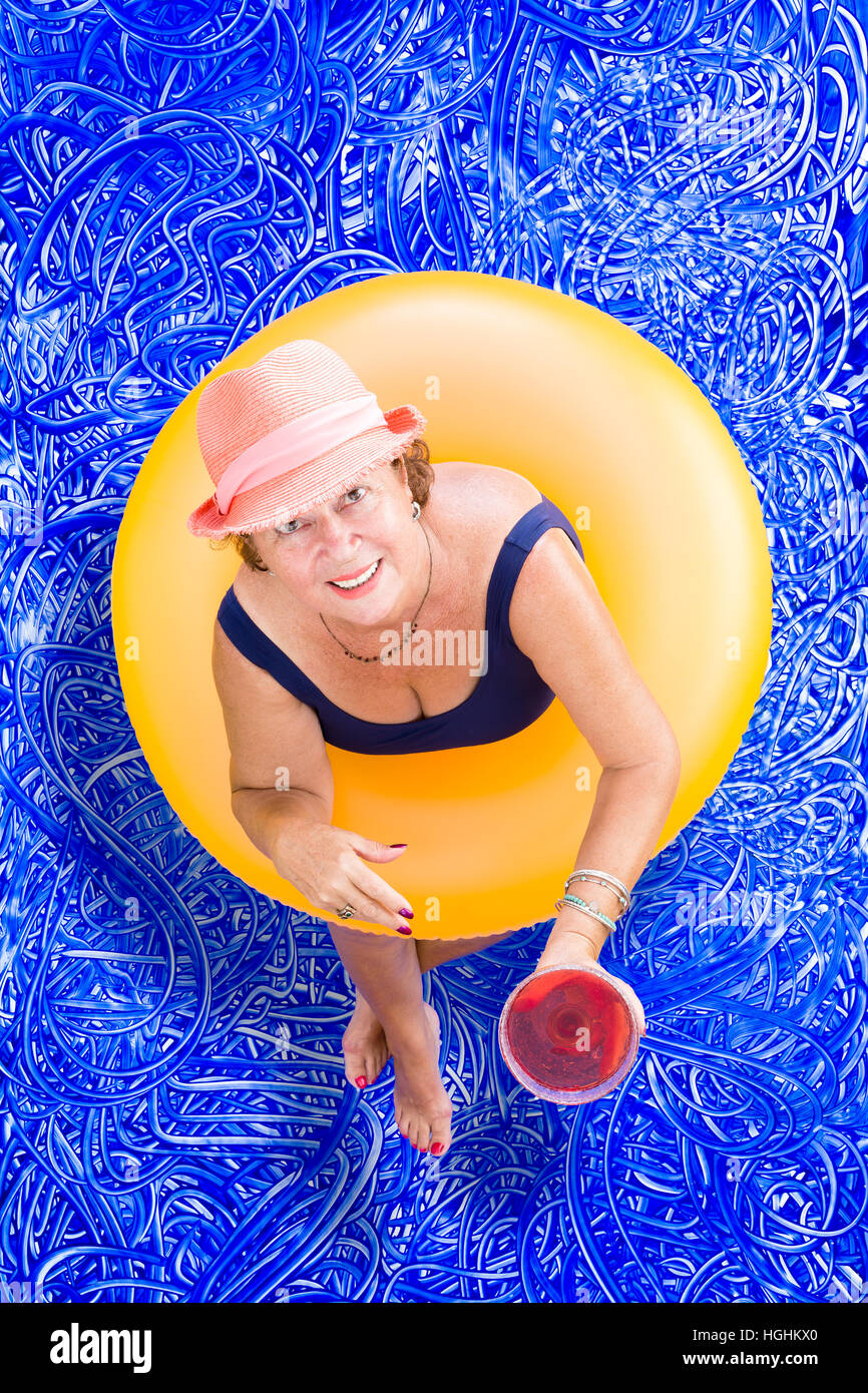 Dame âgée de boire une pina colada cocktail dans la piscine tout en flottant dans un tube en plastique jaune, et l'élever son verre en un toast ou greeti Banque D'Images