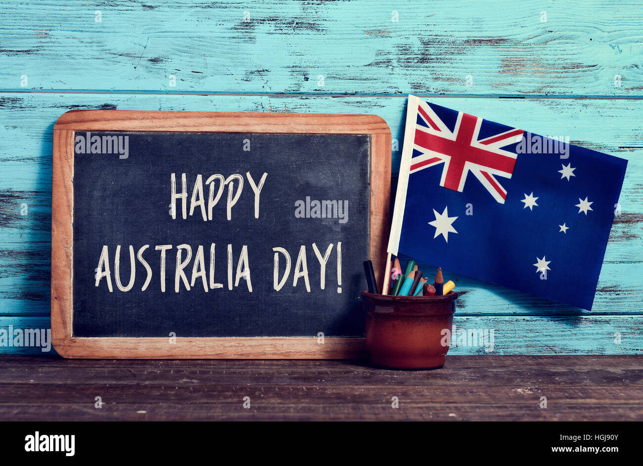 Un tableau sur le texte happy Australia Day écrit en C, un pot à crayons et le drapeau de l'Australie, sur une surface en bois, contre un bleu rustique Banque D'Images