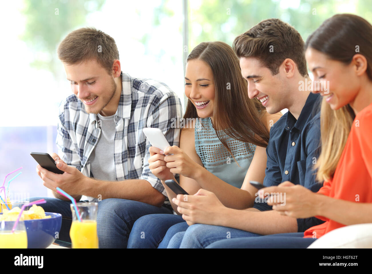 Quatre amis heureux en utilisant leur téléphone portable assis sur un canapé à la maison avec une fenêtre à l'arrière-plan Banque D'Images