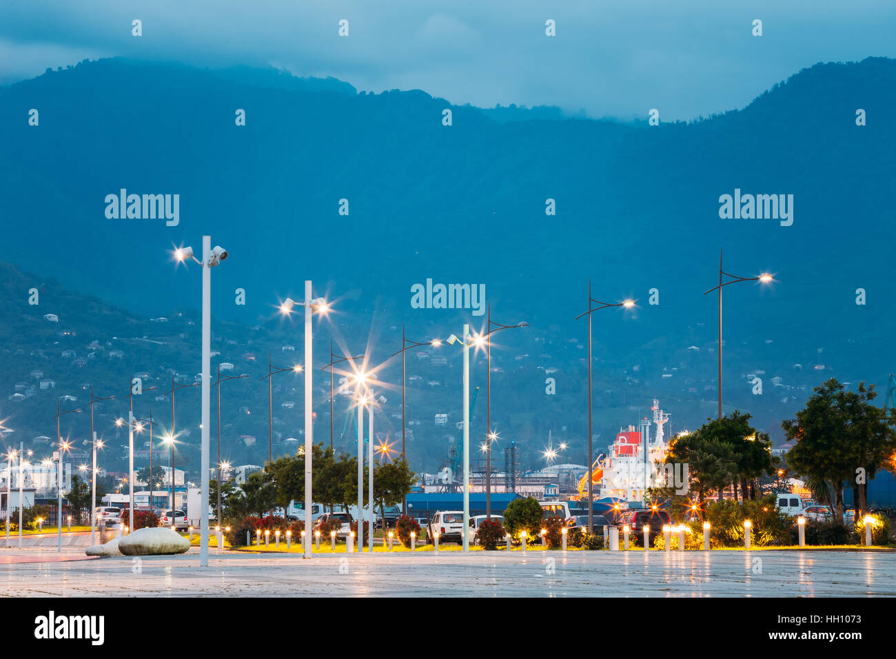 Batumi, Géorgie, l'Adjarie. Lignes de passage de la lumineuse lampadaires au bord de mer à quai dans le parc de miracle. La vue sur la montagne dans la brume du soir. Banque D'Images