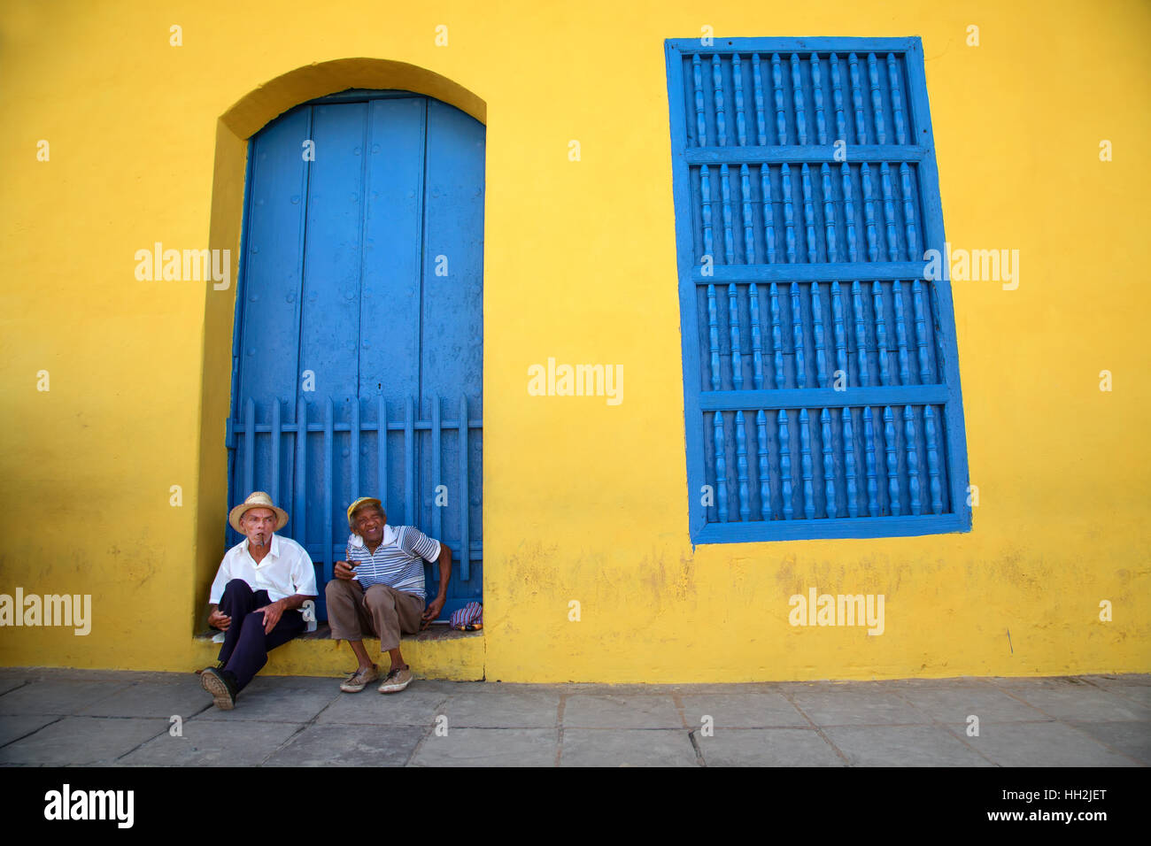Deux hommes sont assis en face d'un mur d'une maison colorée à Santiago de Cuba avec un cigare Cohiba Banque D'Images