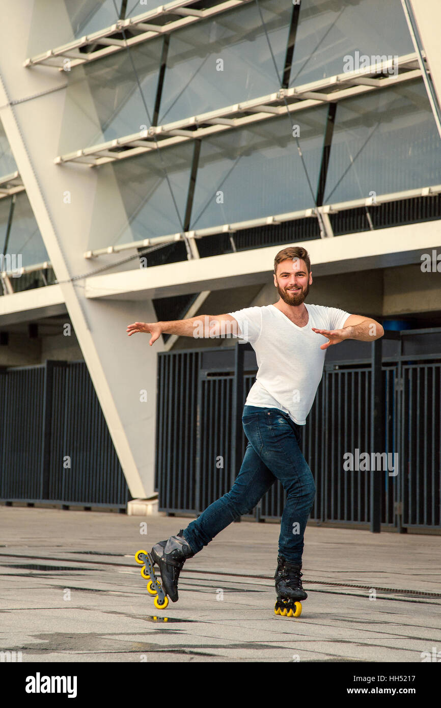 Homme barbu sur rollers debout dans l'arrière-plan de construction. Jeune homme en blanc avec des t-shirts et jeans sur patins à cheval Banque D'Images