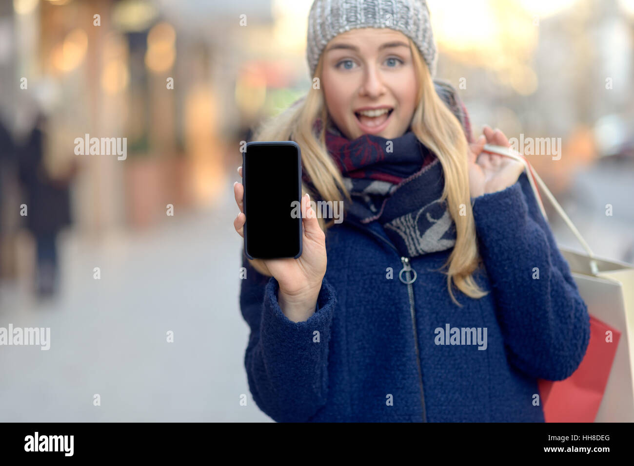 Pretty young woman holding up son téléphone mobile avec un écran blanc comme elle se tient sur une rue urbaine en hiver chaudement vêtu d'un tricot de fils de sca Banque D'Images