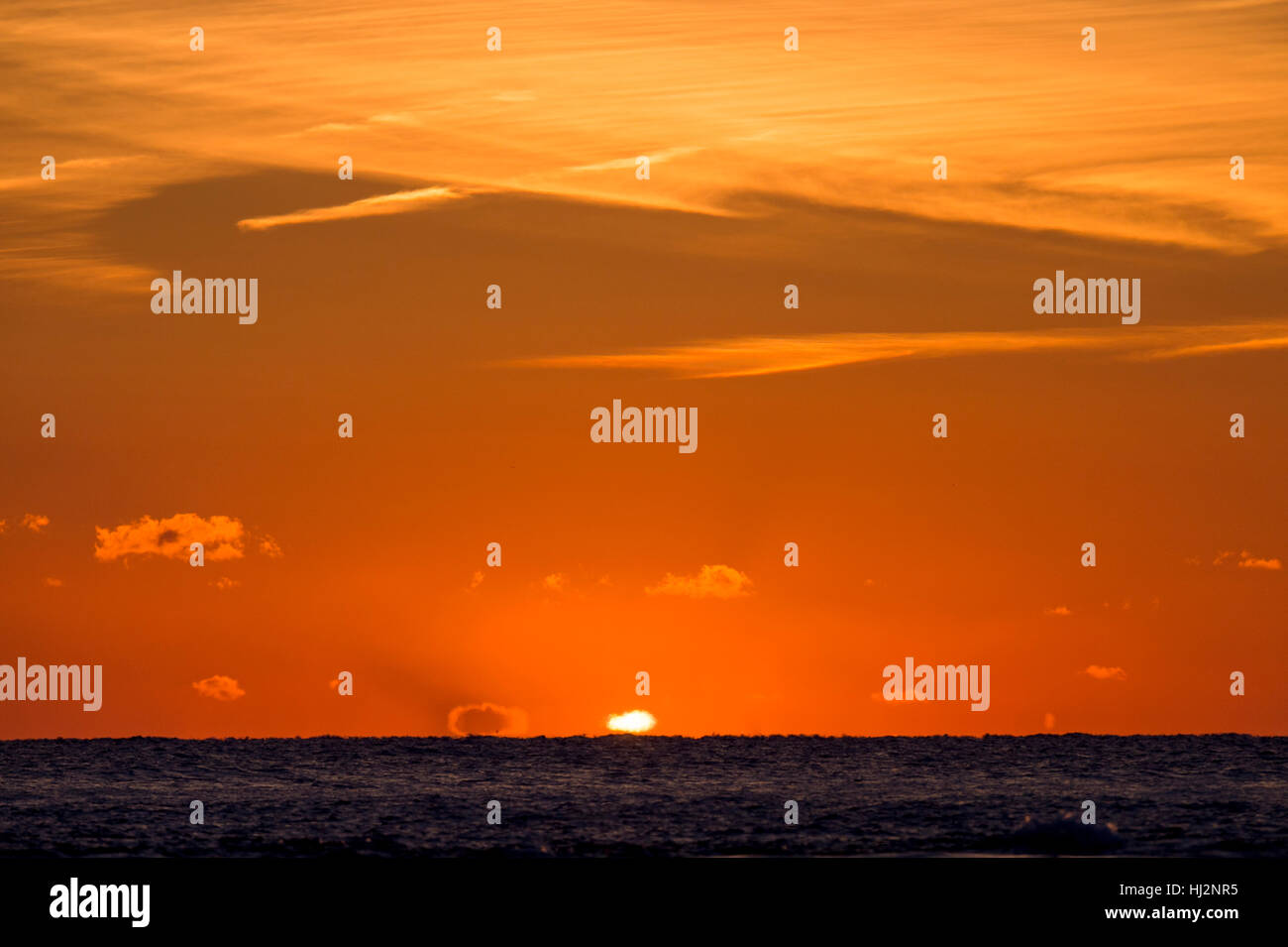 Le soleil peut à peine être vu lorsqu'il s'ouvre sur l'océan avec de minces nuages dans le ciel orange vif. Banque D'Images