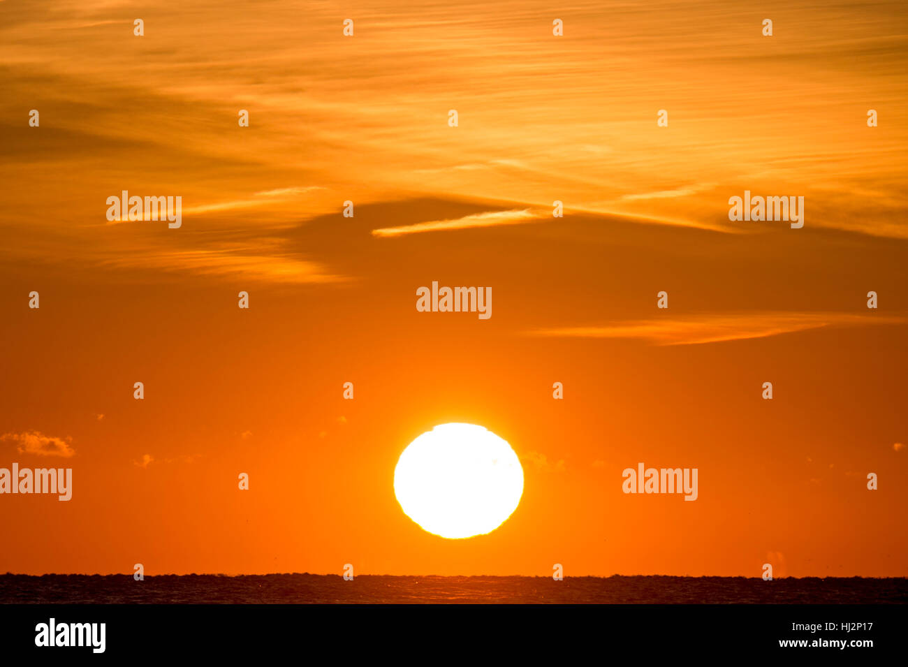 Un grand soleil jaune se couche sur l'océan avec de minces nuages dans le ciel orange. Banque D'Images