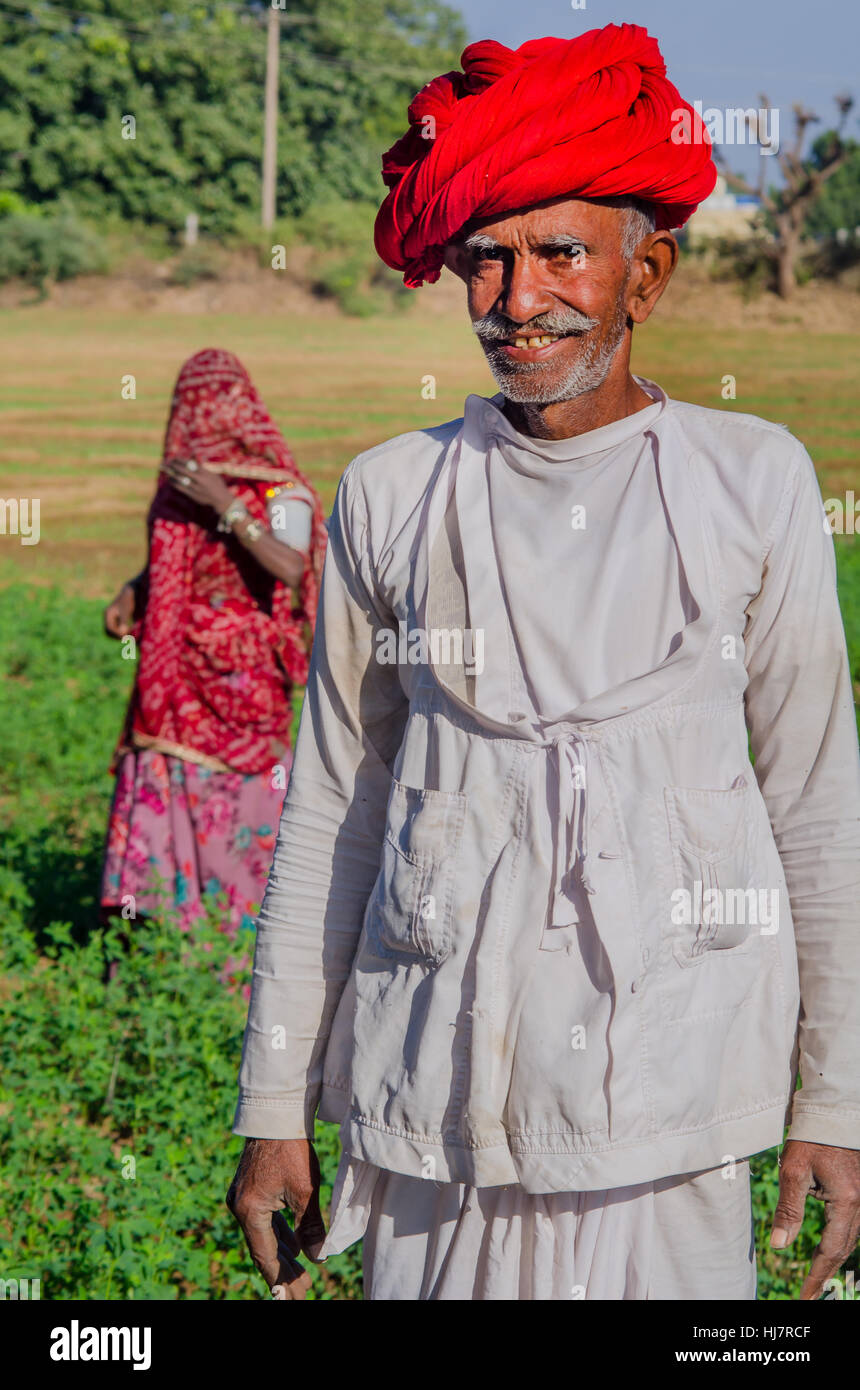 Râjasthânî Senior citizen wearing red turban dans sa ferme avec sa femme en arrière-plan. Banque D'Images