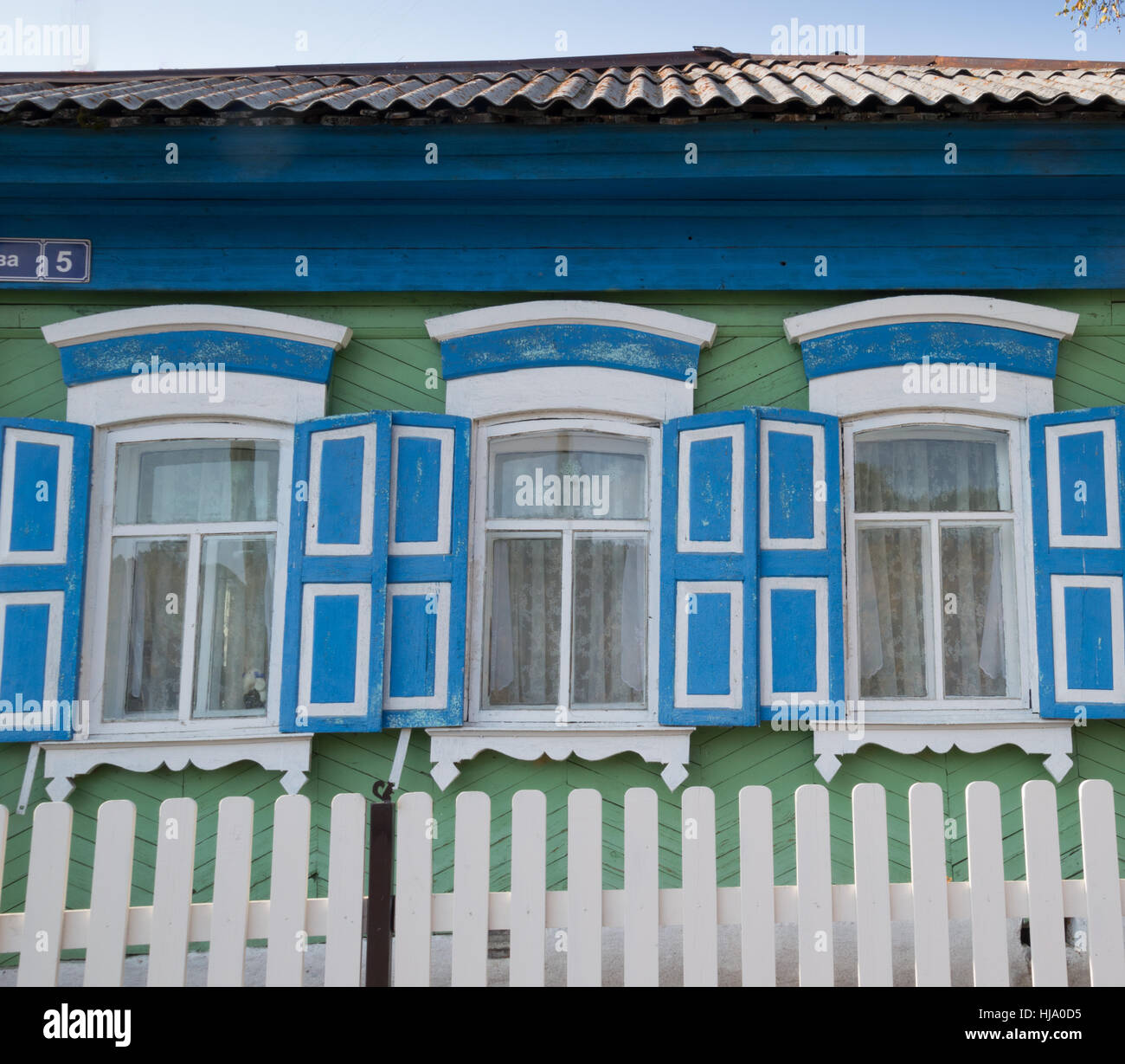 Une maison verte avec garniture de fenêtre blanc et bleu et des volets roulants. Les fenêtres ont des rideaux de dentelle. Une clôture blanche est en face Banque D'Images