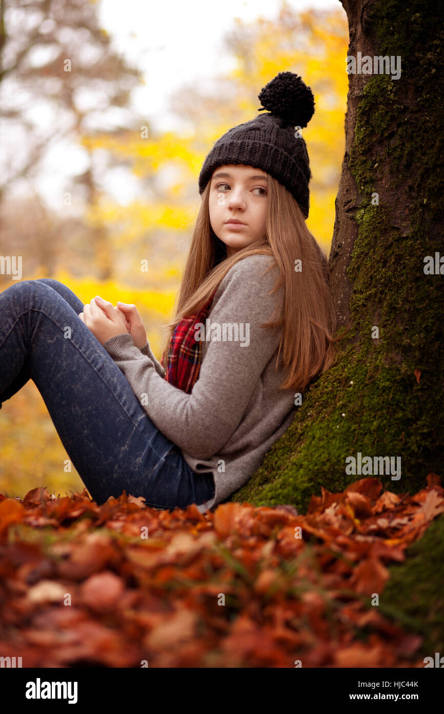 Jolie jeune fille assise à côté d'un arbre en automne avec un tapis si les feuilles sur le sol Banque D'Images