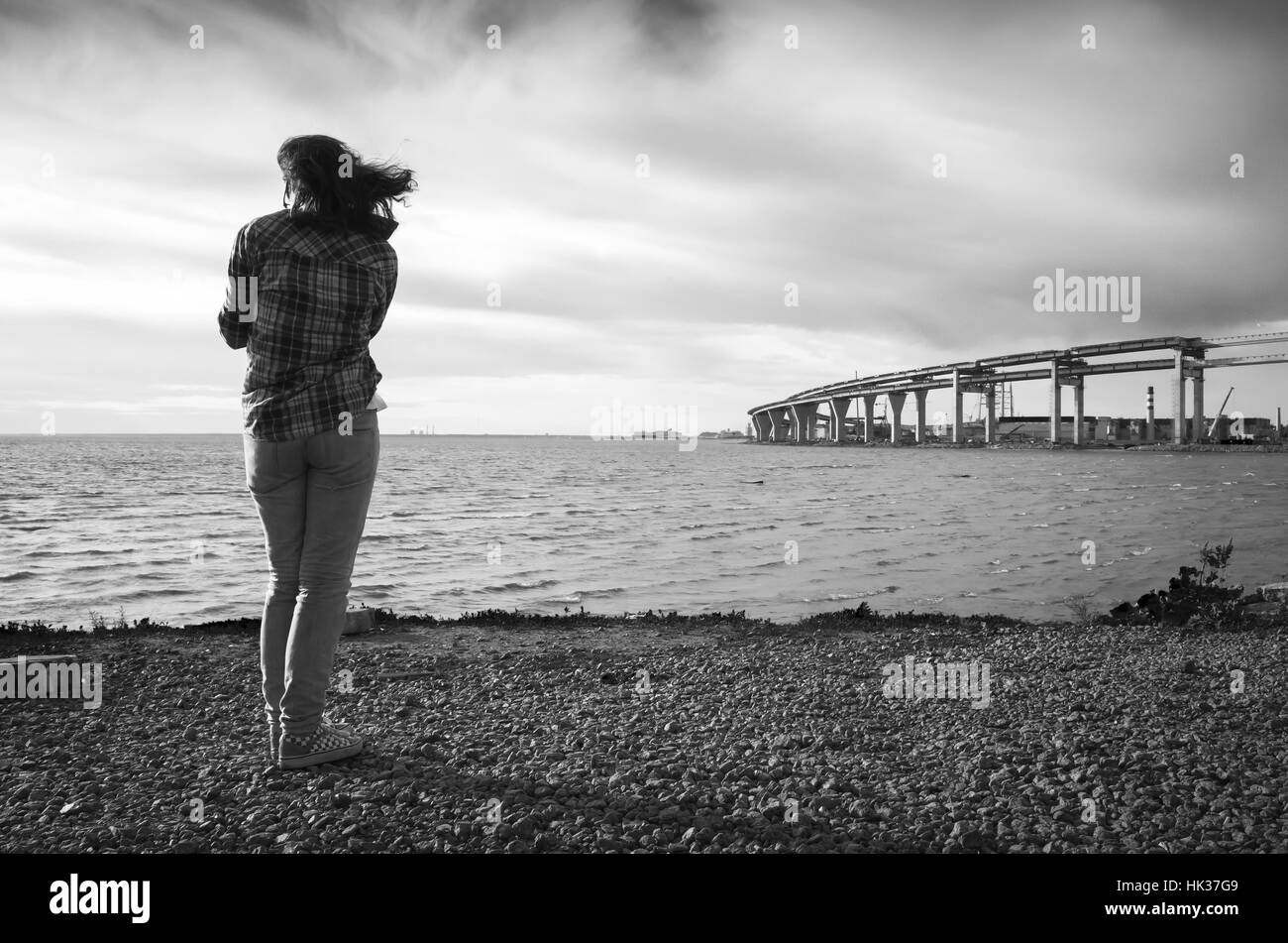 Jeune fille se tient sur un littoral, pont moderne en construction sur un horizon, photo en noir et blanc Banque D'Images