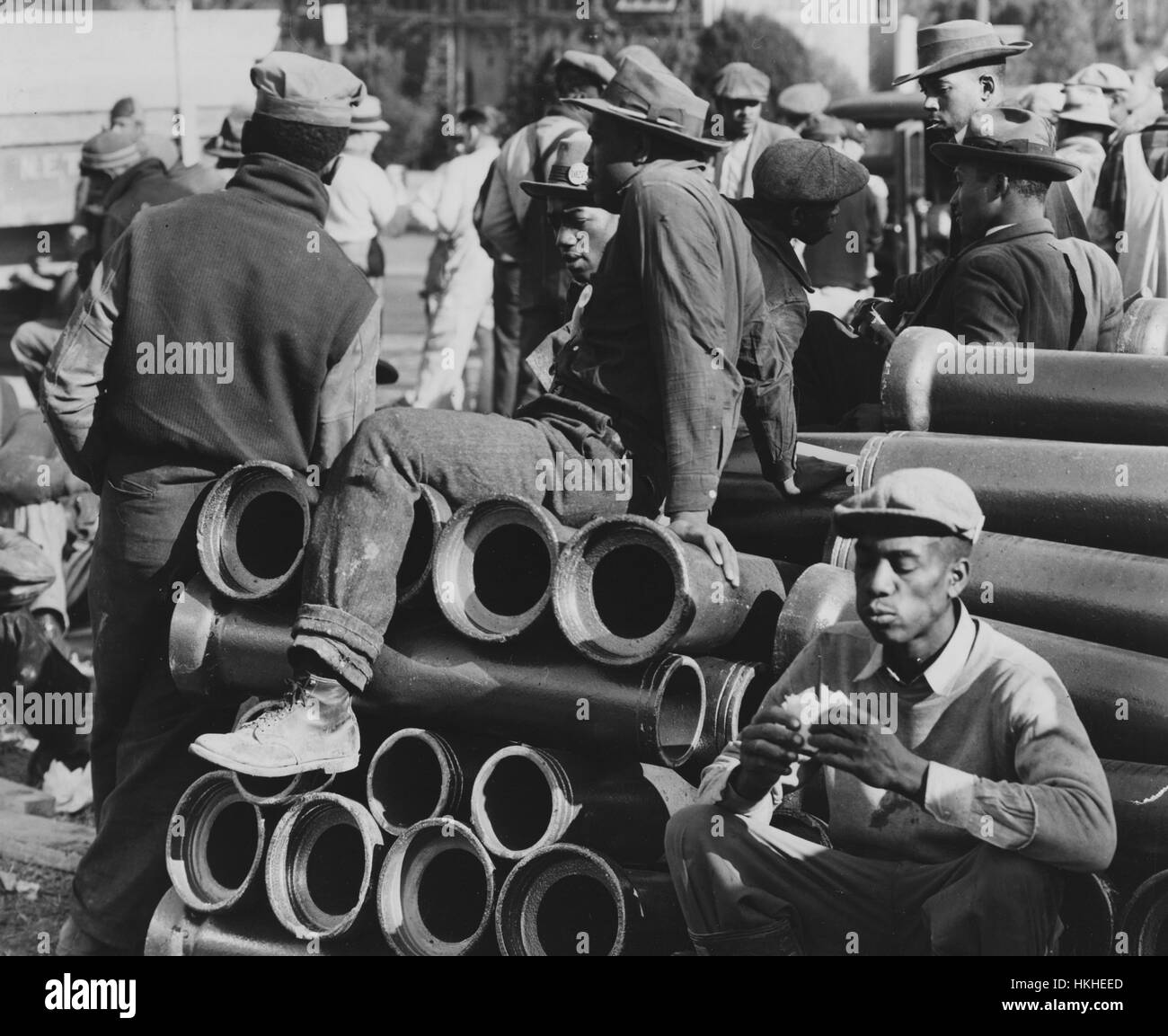 Photographie en noir et blanc d'un groupe de travailleurs afro-américains pour un bureau de la défense d'urgence, une guerre d'urgence fédérale des États-Unis agence créée pour coordonner les mesures fédérales et d'état pour la protection des civils en cas de guerre, la construction de logements d'urgence, travail pendant la pause déjeuner, debout autour et assis sur de grands tuyaux, Washington, DC, décembre, 1941. À partir de la Bibliothèque publique de New York. Banque D'Images