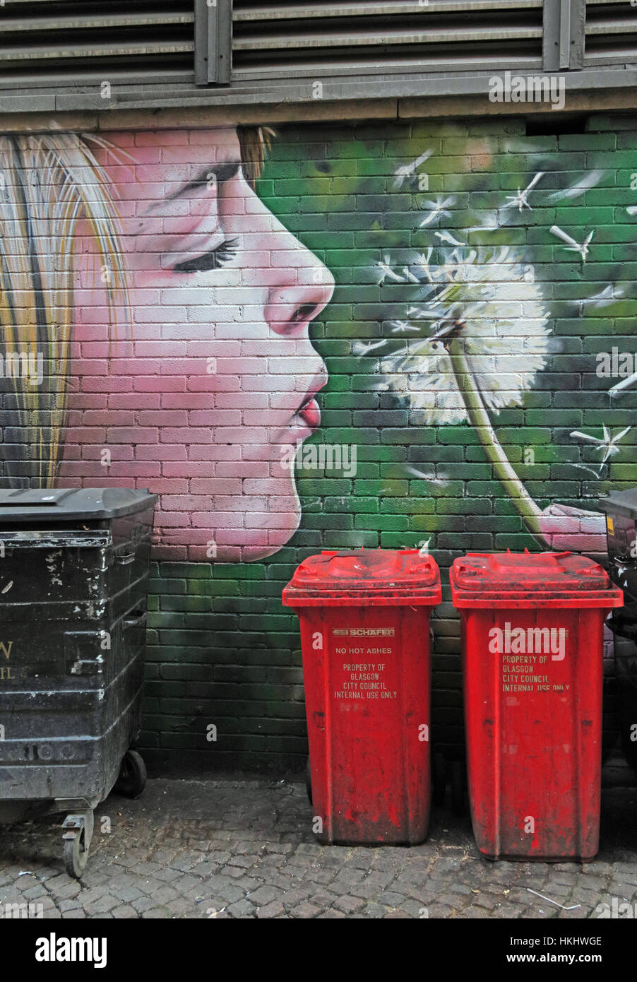 Femme blonde soufflant les graines d'une horloge pissenlit, au-dessus des poubelles, Glasgow, Écosse, Royaume-Uni Banque D'Images
