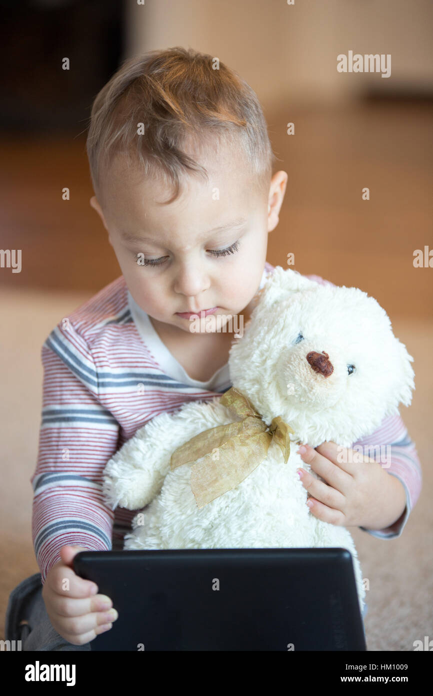 Little toddler boy holding a teddy bear tout en jouant avec l'ordinateur tablette. Banque D'Images