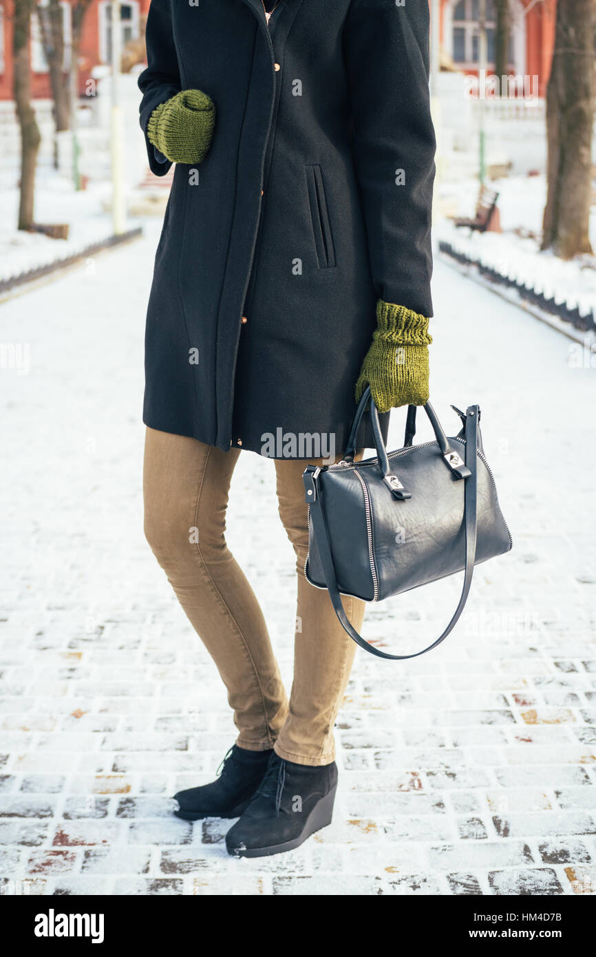 Jeune femme mince dans un manteau noir et des gants verts et un sac à main  tenant debout sur le trottoir enneigé Photo Stock - Alamy