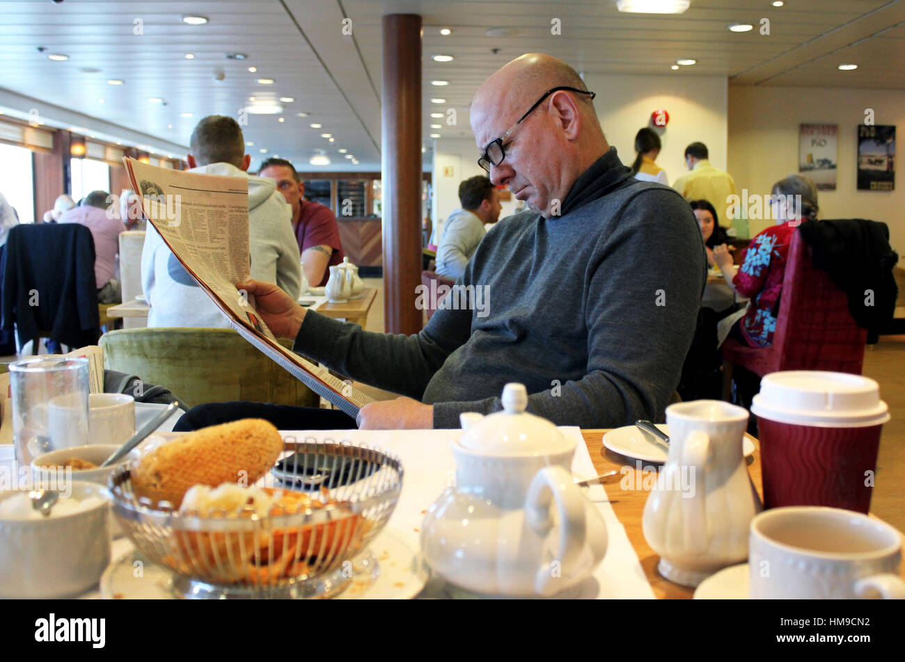 Middle aged man reading newspaper tout en prenant le petit déjeuner à bord d'un ferry. Banque D'Images