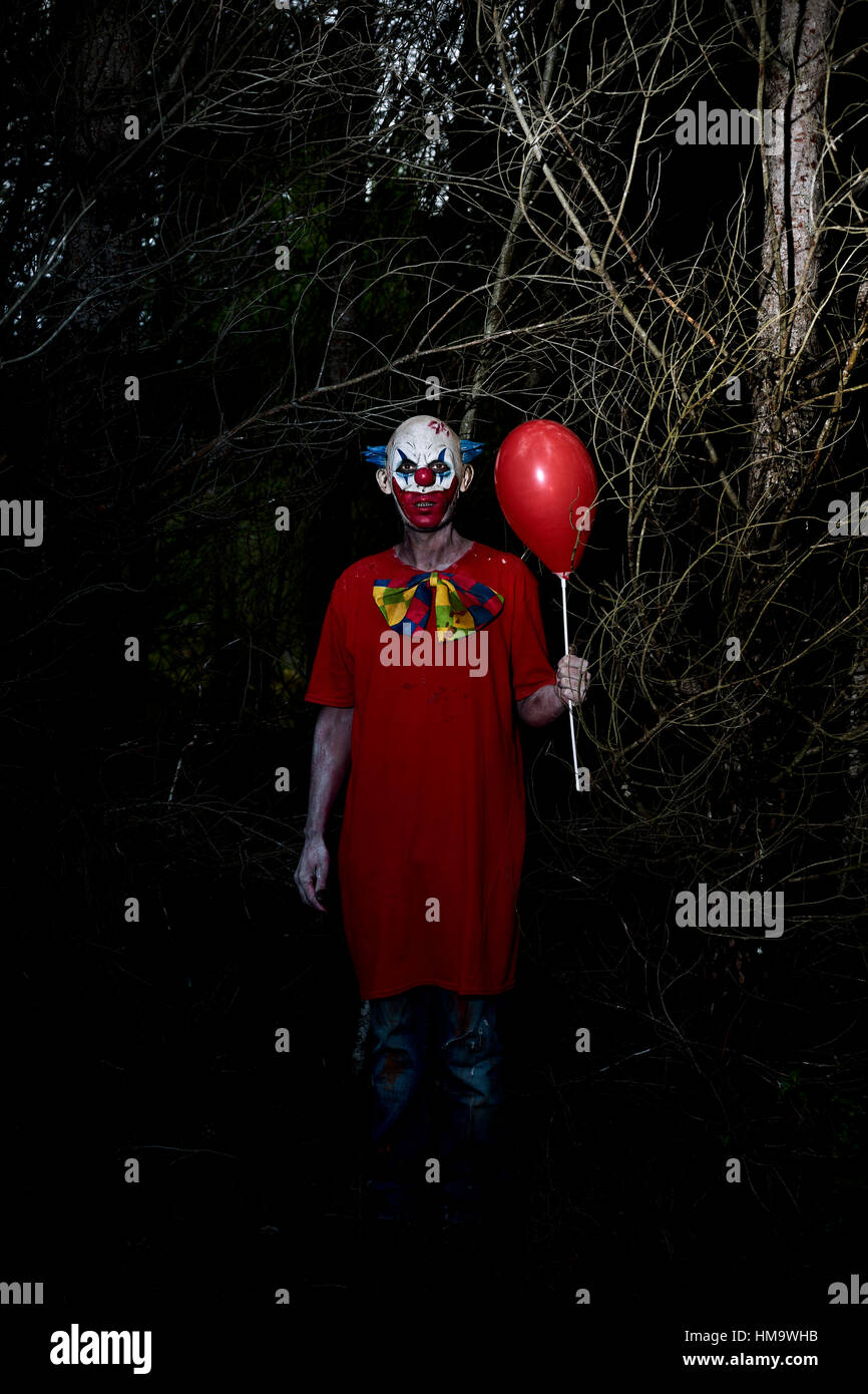 Un mal effrayant clown portant un costume sale, tenant un ballon rouge dans sa main, dans les bois La nuit Banque D'Images