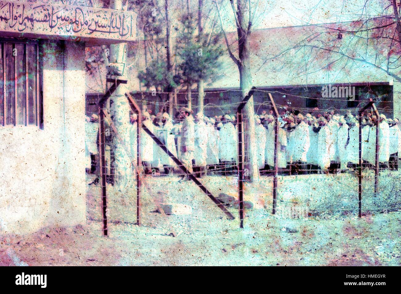 Vue à travers une clôture de barbelés d'un groupe d'hommes vêtus de robes blanches et de bandeaux, debout avec les poings soulevées dans une grande cour en Iran, mars, 1983. Un signe en partie visible dans le coin supérieur gauche affiche l'avant-plan d'un proverbe écrit en arabe à partir de l'Al Tawbah P-111 Adoption du Coran. La partie visible se traduit par : "En fait les croyants ont échangé leurs vies et leurs biens au Dieu pour [un morceau de ciel.].'. D'importants dégâts sur l'original. (Photo de la Collection Morse/Gado/Getty Images). Banque D'Images