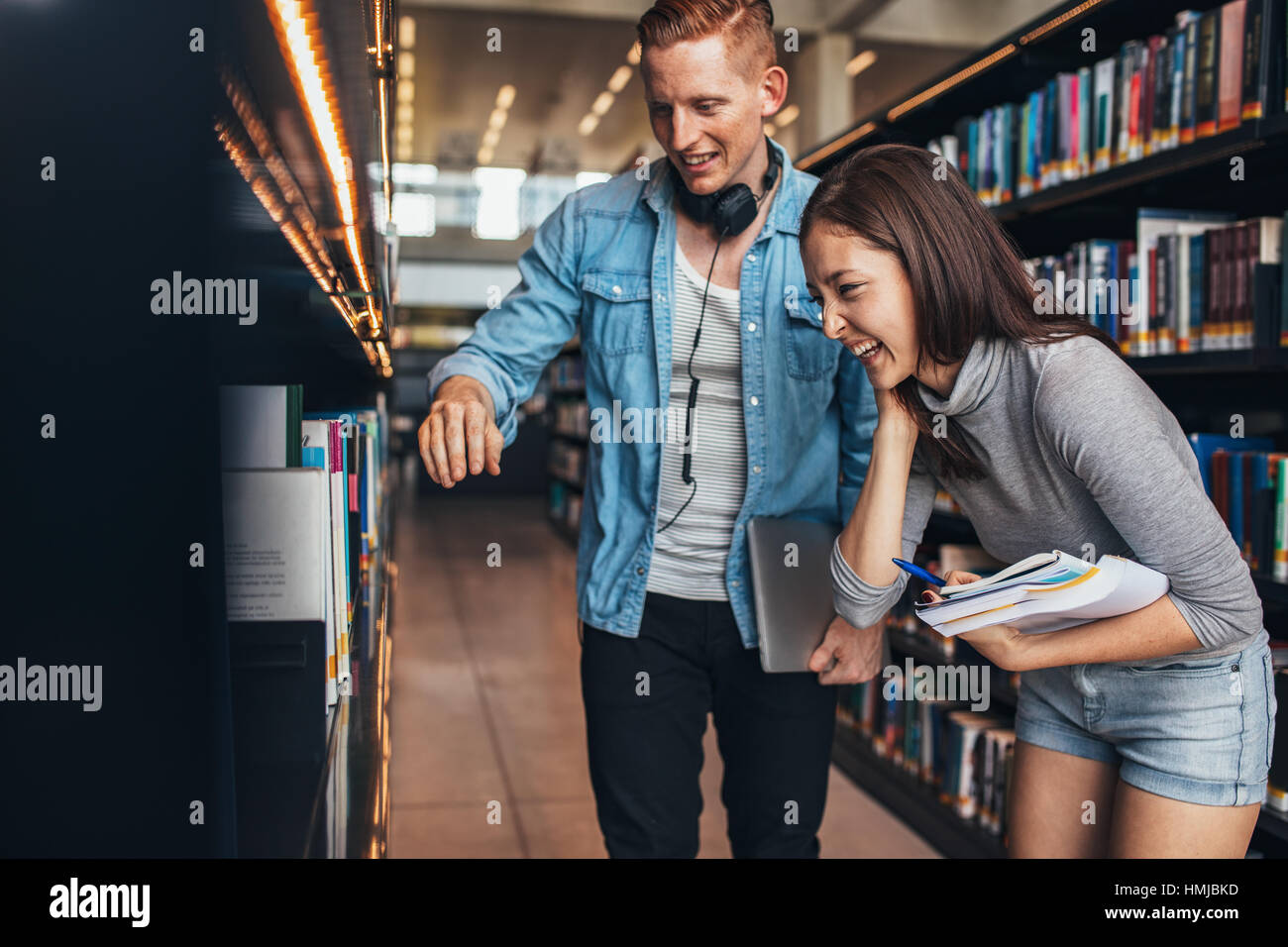 Image of young man and woman standing by book shelf en bibliothèque et à la recherche de livres. Les élèves de collège à la bibliothèque pour les livres. Banque D'Images