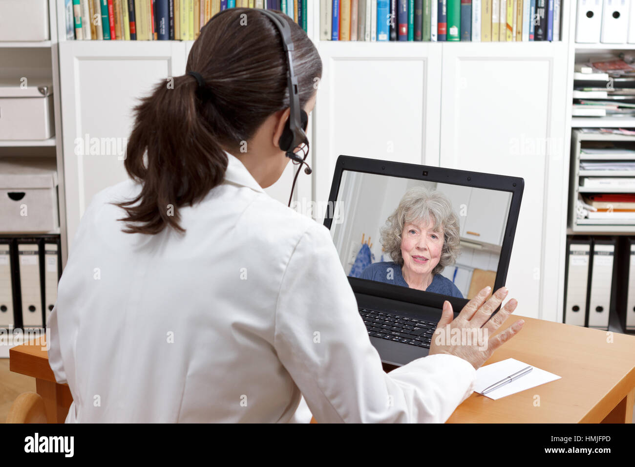 Femme médecin de la gériatrie dans son bureau de la chirurgie avec casque en face de son ordinateur portable parlant via appel vidéo avec une vieille patiente sur son prescrit Banque D'Images