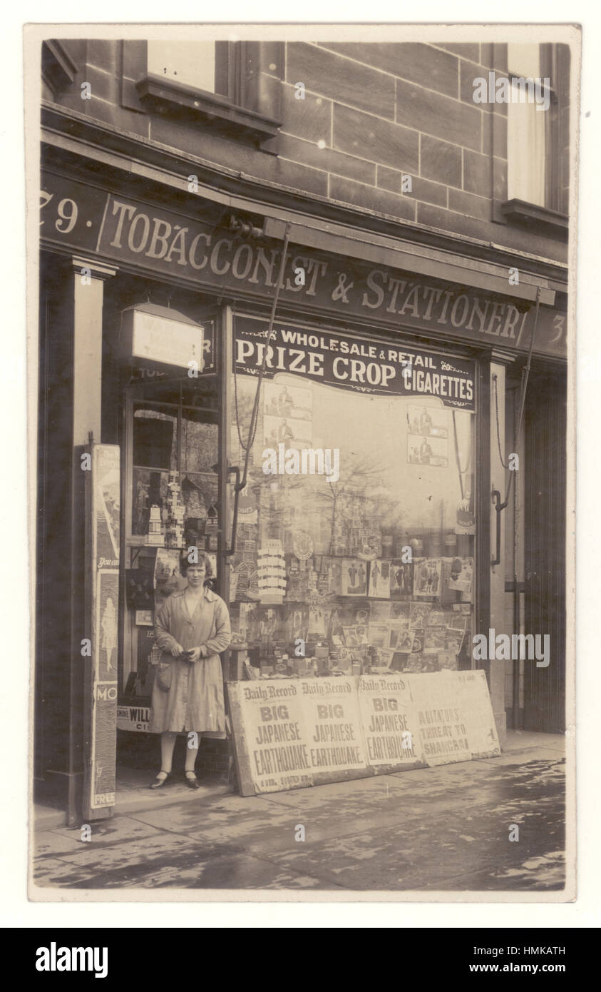 Carte postale de femme titulaire / propriétaire / shop keeper ou du personnel en dehors de ses assistants /tabac et des fournitures de boutique, qui est également un des marchands. Nouvelles d'un grand séisme japonais sur un panneau à l'extérieur de la boutique date probablement la photo de 1923, les produits affichés dans la fenêtre de devantures, UK Banque D'Images