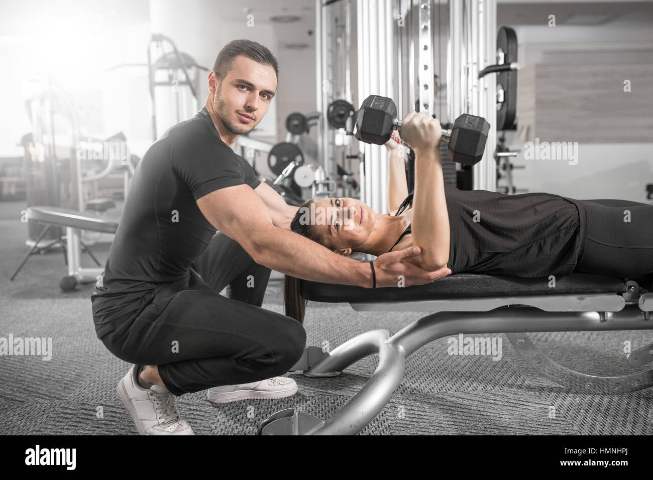 Remise en forme des jeunes adultes couple. Femme poitrine et épaule dumbell press sur banc avec entraîneur personnel l'aider et donner des instructions. Banque D'Images