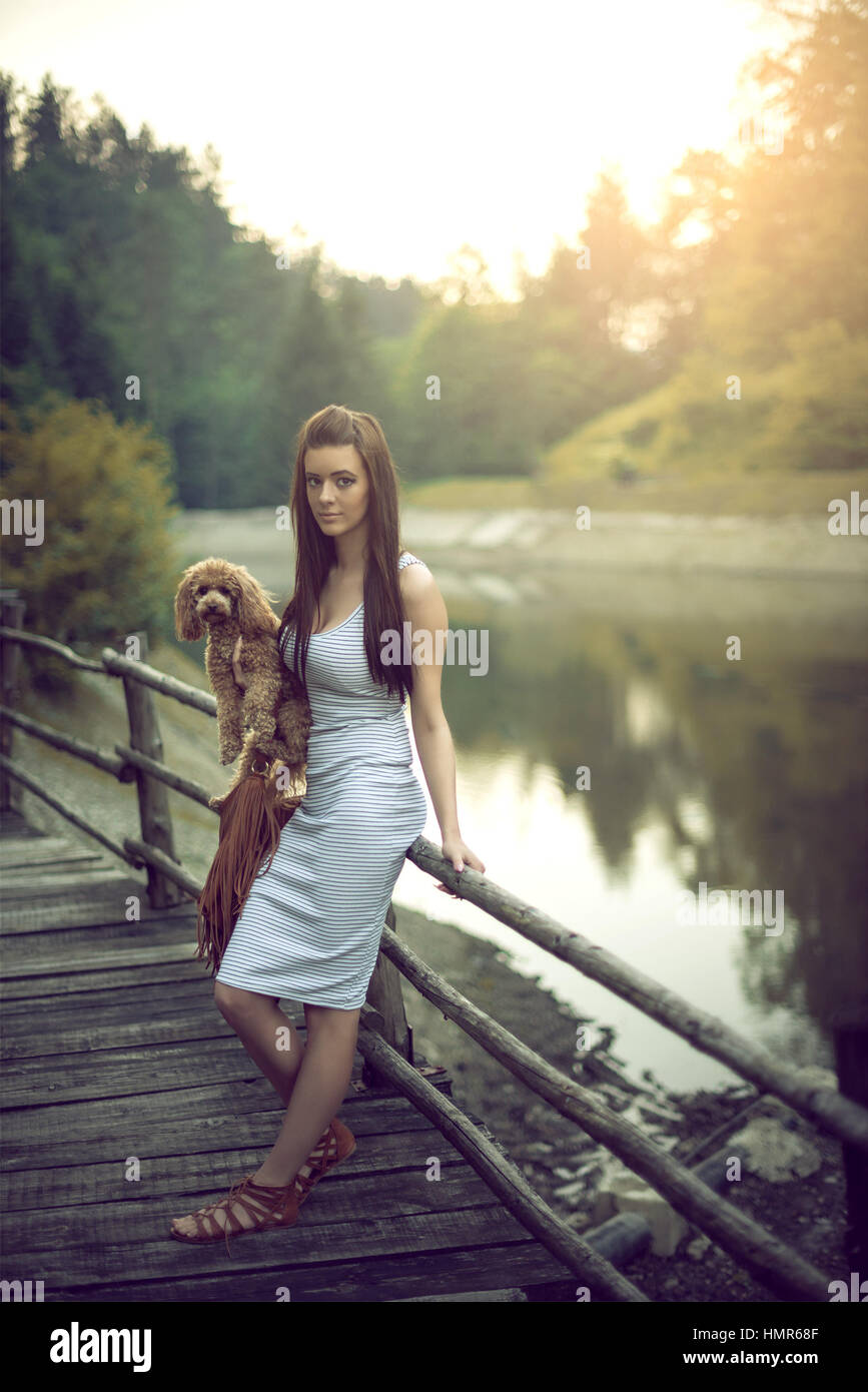 Belle jeune femme brune adultes posant au lac de montagne avec son jouet chien caniche rouge. La tonalité de l'image. Filtres artistiques appliquées. Banque D'Images