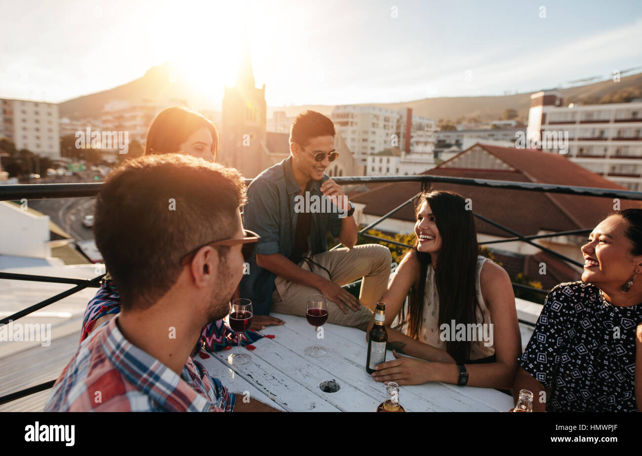 Groupe d'amis ayant une partie sur le toit. Les jeunes hommes et les femmes assis autour d'une table avec des boissons et de rire. Banque D'Images