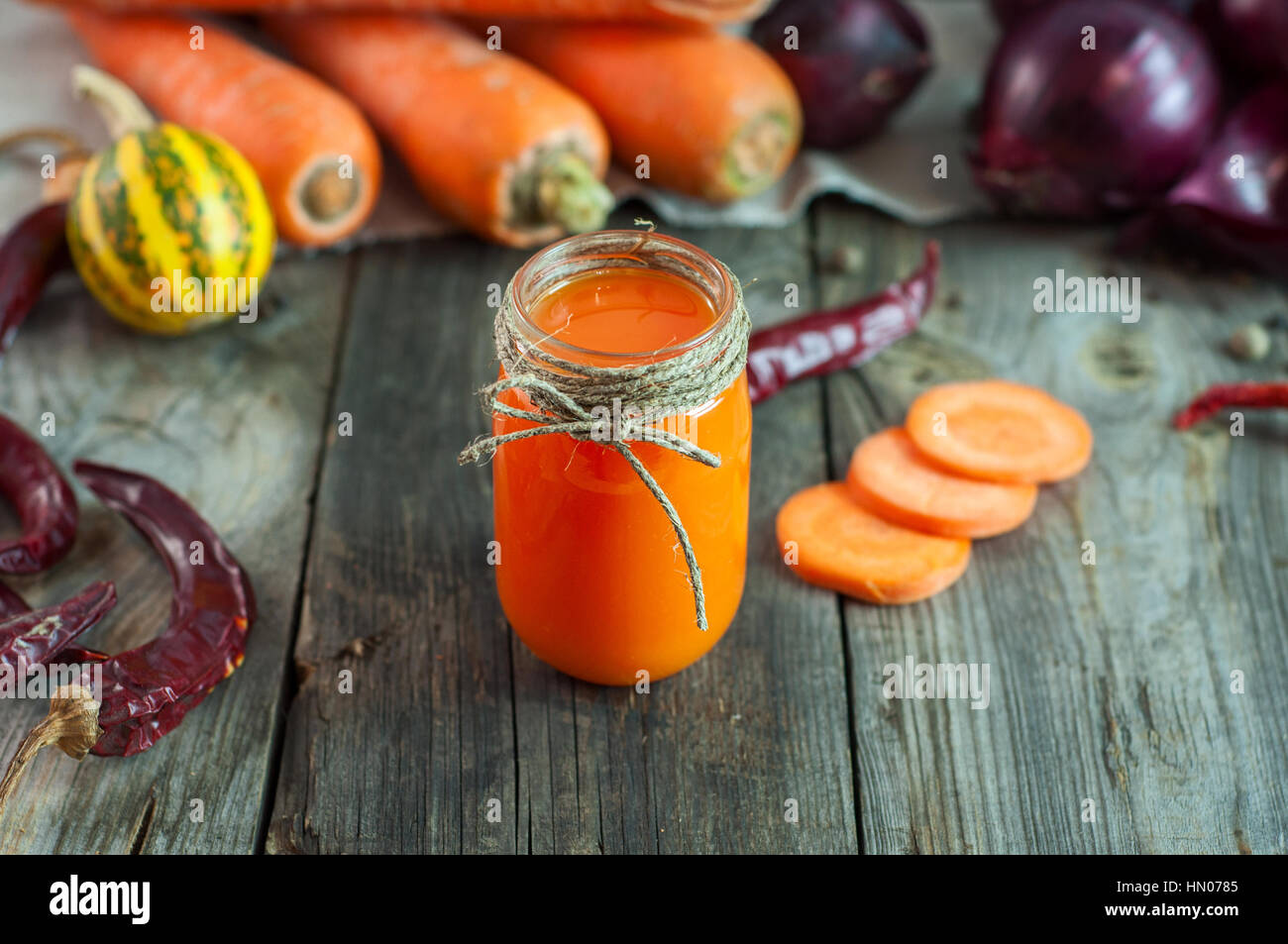 Le jus de carotte dans un petit pot transparent entre les légumes frais, sur une surface en bois gris Banque D'Images