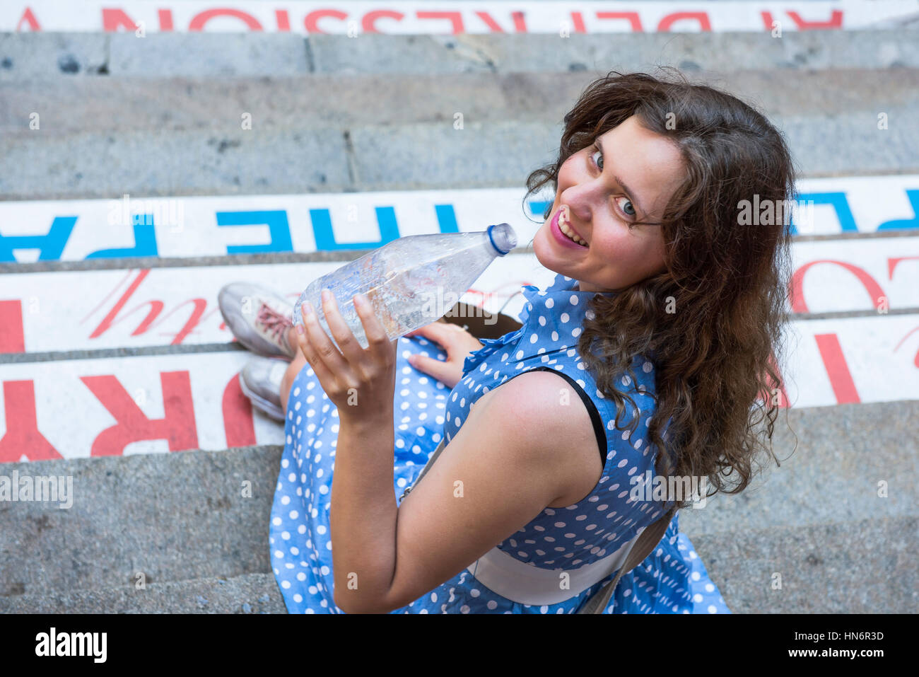 Jeune femme avec robe à pois bleu assis sur les marches la consommation de boissons de l'eau dans une bouteille transparente et de rire Banque D'Images