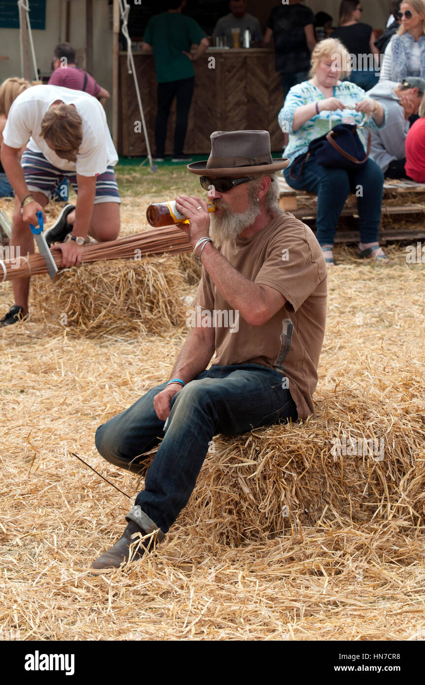 Homme avec barbe et hat sitting on bale of hay de boire une bouteille de bière au Port Eliot Cornwall Festival Banque D'Images