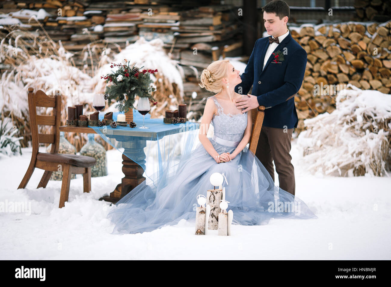 Mariage d'hiver sur la neige en plein air et le bois de l'arrière-plan. Mariée est assise, le marié se tient à proximité. À côté d'eux est le tableau avec des gobelets de vin et bouquet. Banque D'Images
