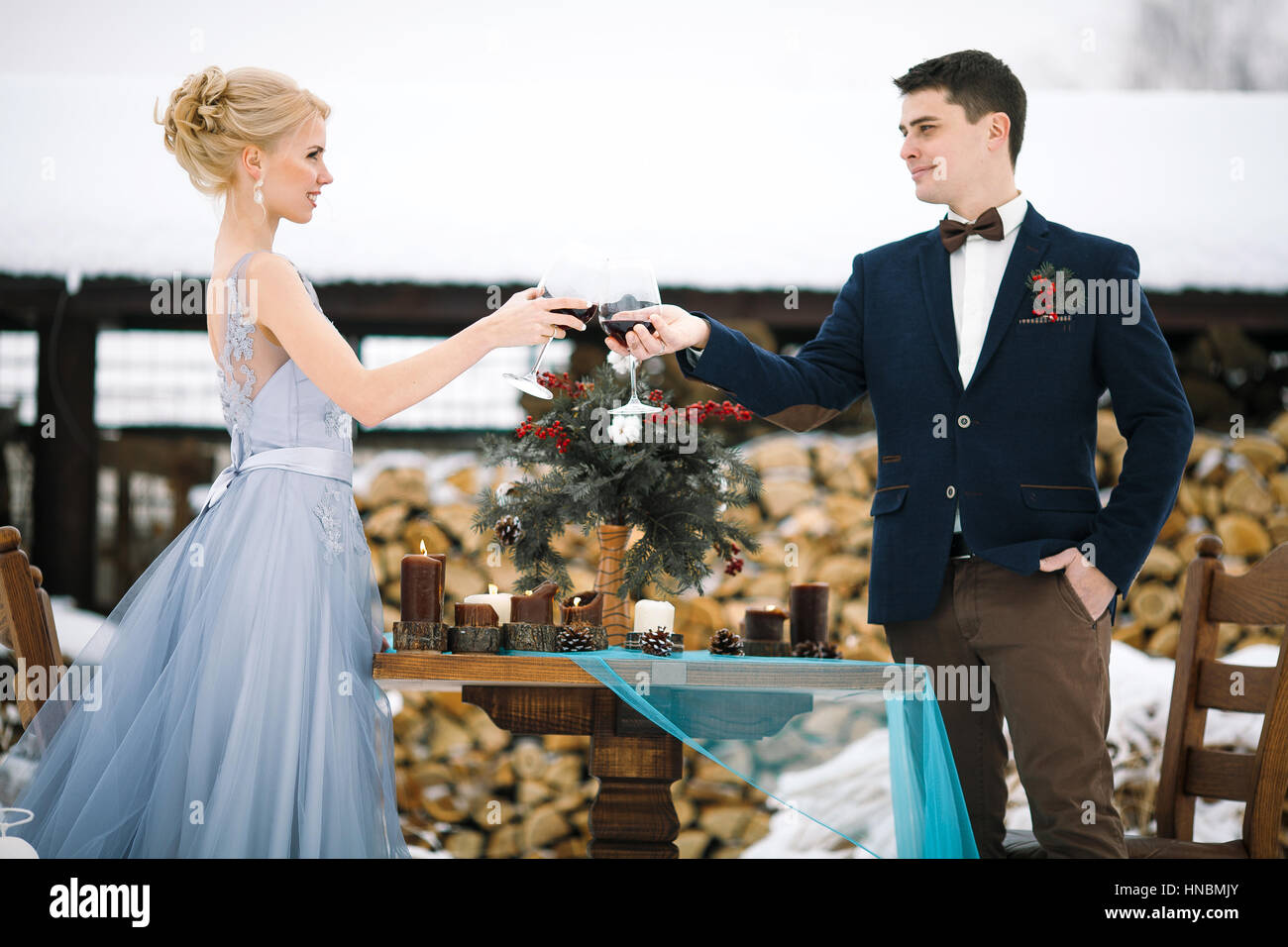 Mariage d'hiver en plein air, sur fond de bois. Jeunes mariés restent debout près de la table et les verres de vin. clink À côté d'eux est le tableau avec bouquet. Banque D'Images