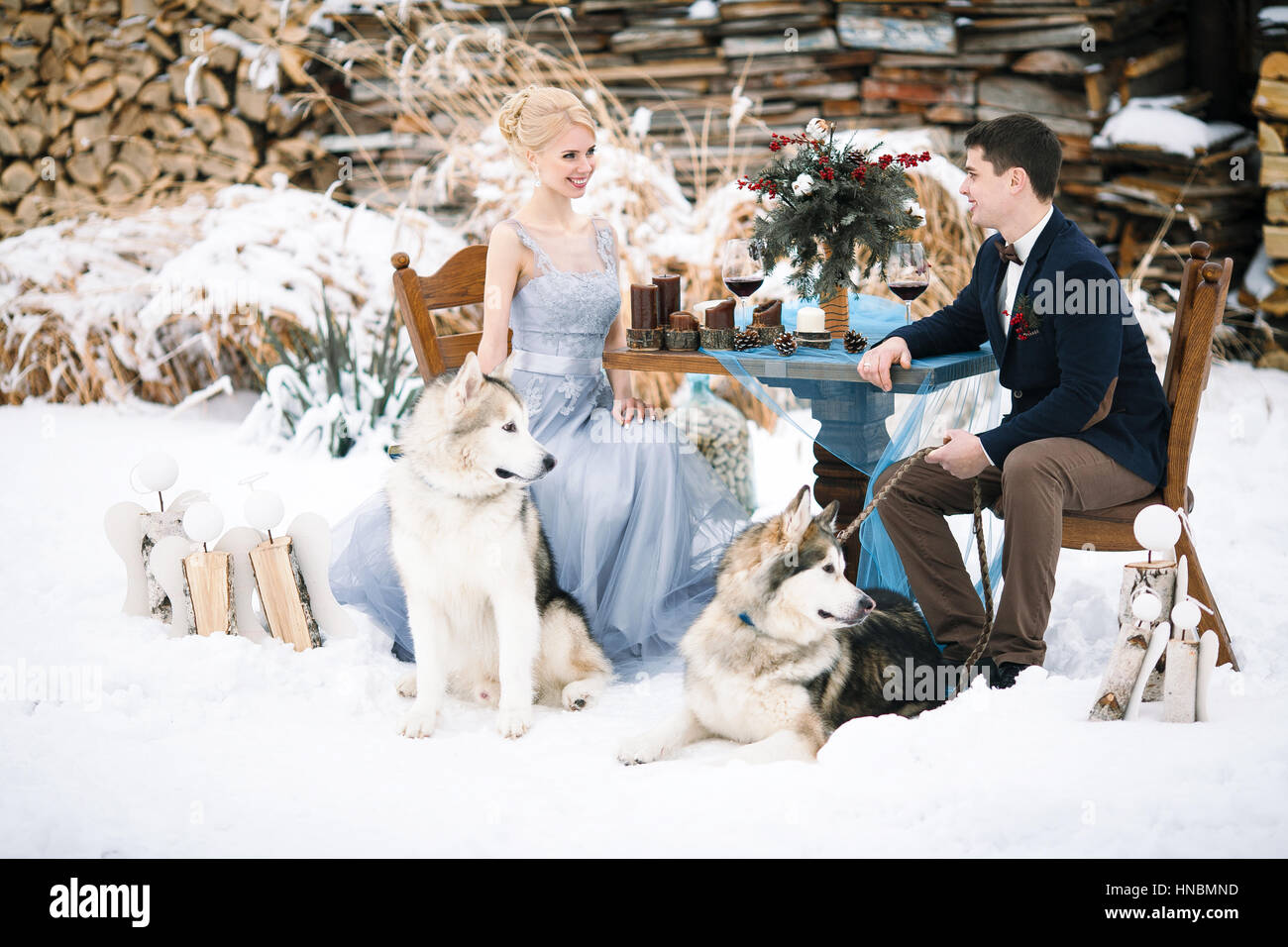 Mariée et le marié en hiver avec deux chiens malamute assis à table. Sur la table sont des verres à vin, des bougies et du bouquet. Banque D'Images