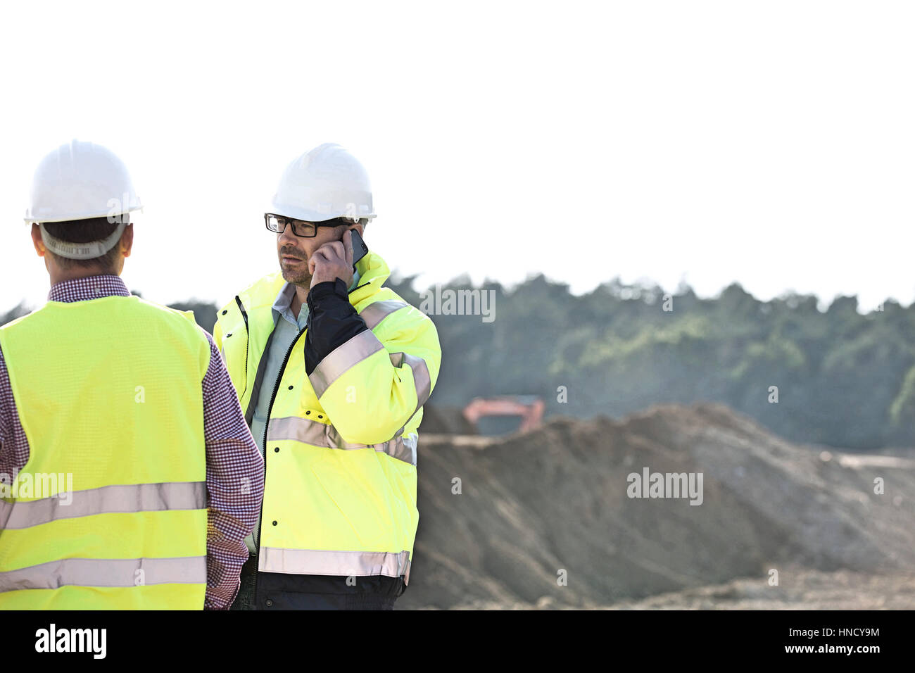 Superviseur en utilisant la téléphonie mobile en position debout avec collègue at construction site contre le ciel clair Banque D'Images