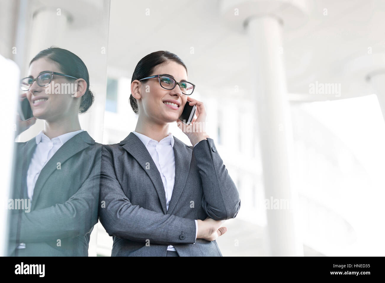 Happy businesswoman using cell phone while leaning on glass wall Banque D'Images