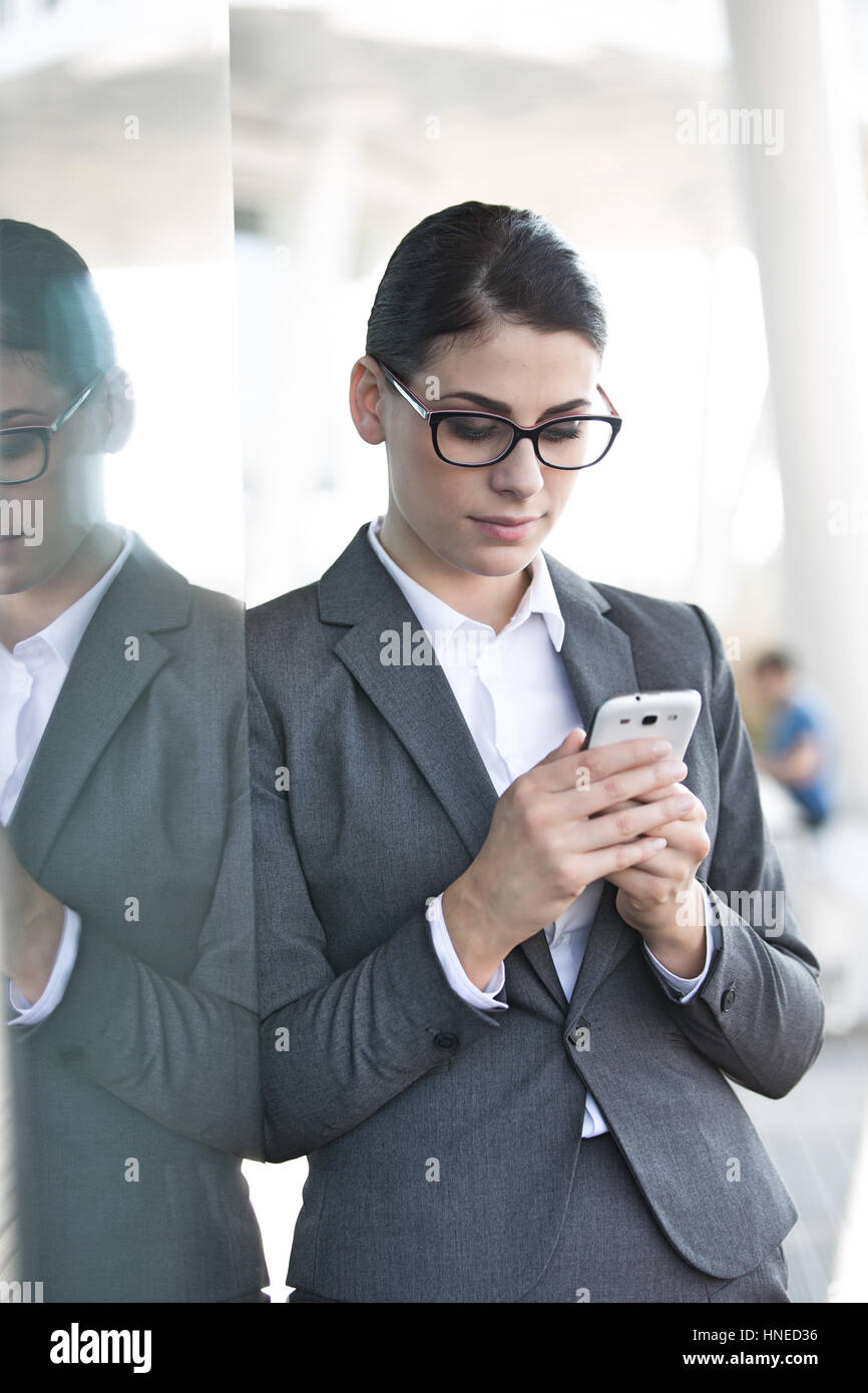 Businesswoman using cell phone tout en s'appuyant sur la paroi de verre Banque D'Images