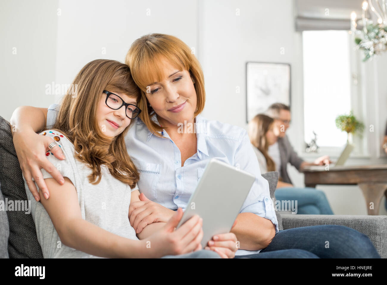 Tendre mère et fille à l'aide de tablette numérique avec la famille assis en arrière-plan à la maison Banque D'Images