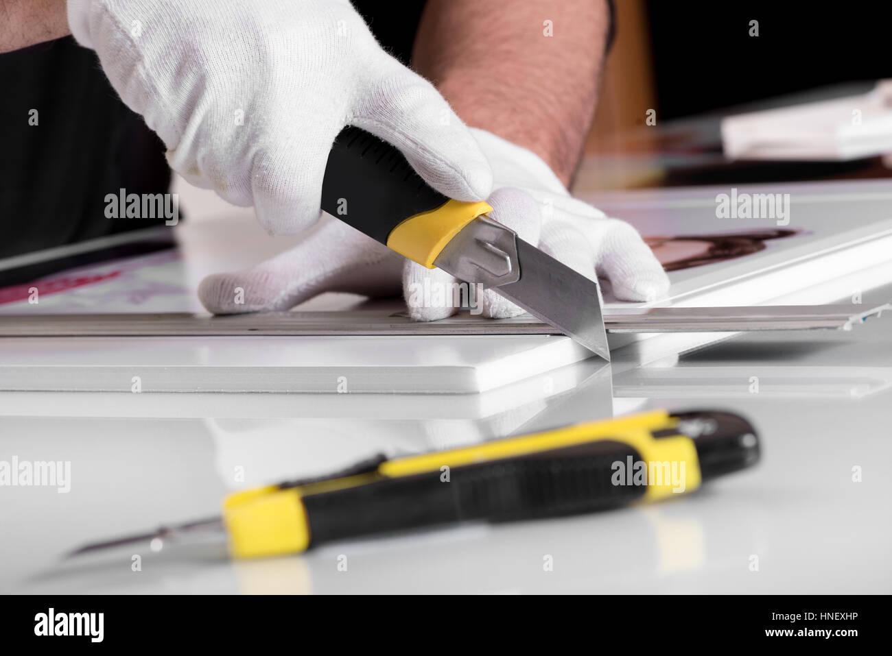Mains Gants professionnel en coupant les bords de la photographie imprimée sur la carte du panneau en PVC blanc Foamex avec un autre couteau de table dans foregroun Banque D'Images
