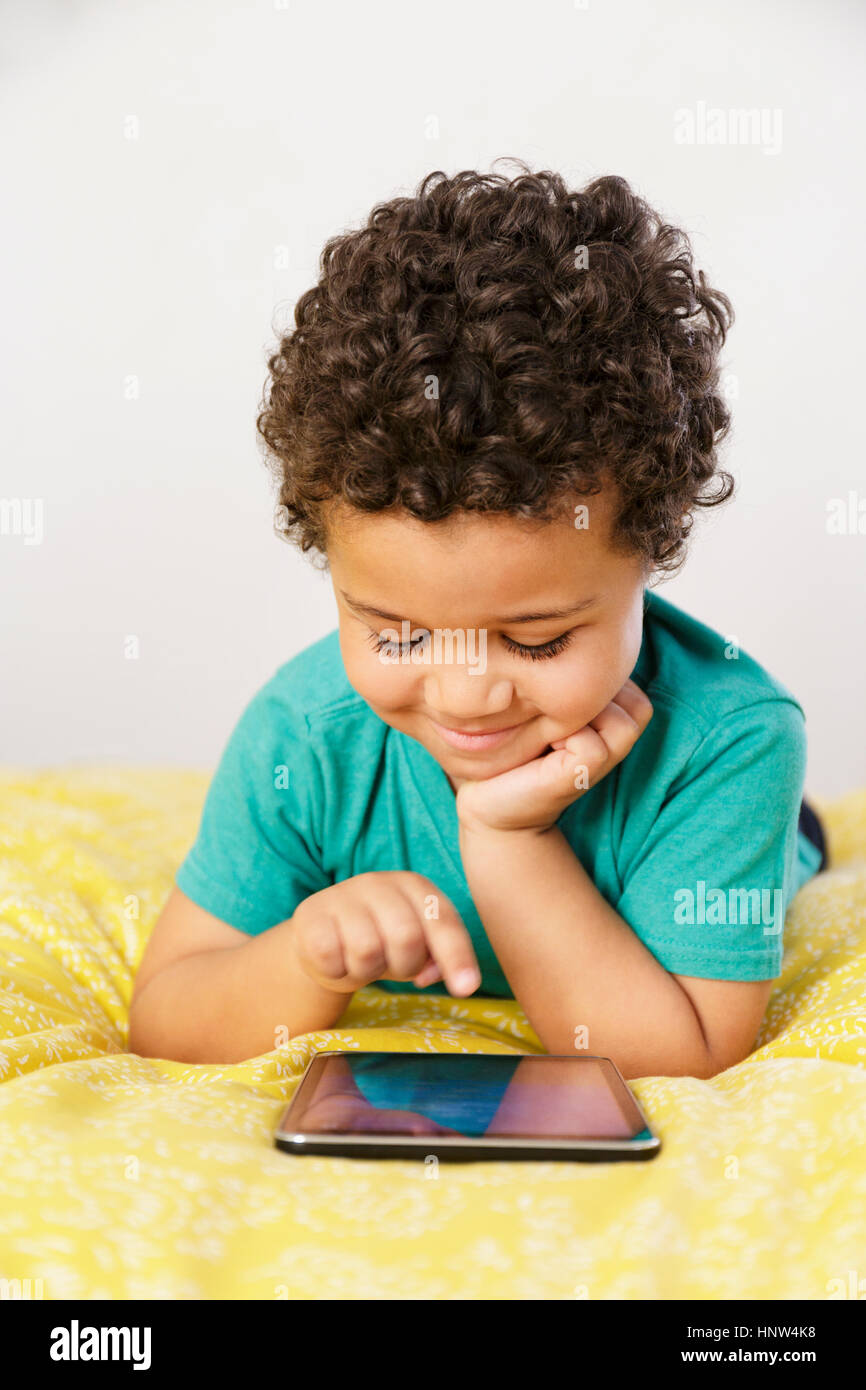 Smiling Mixed Race boy laying on bed using digital tablet Banque D'Images