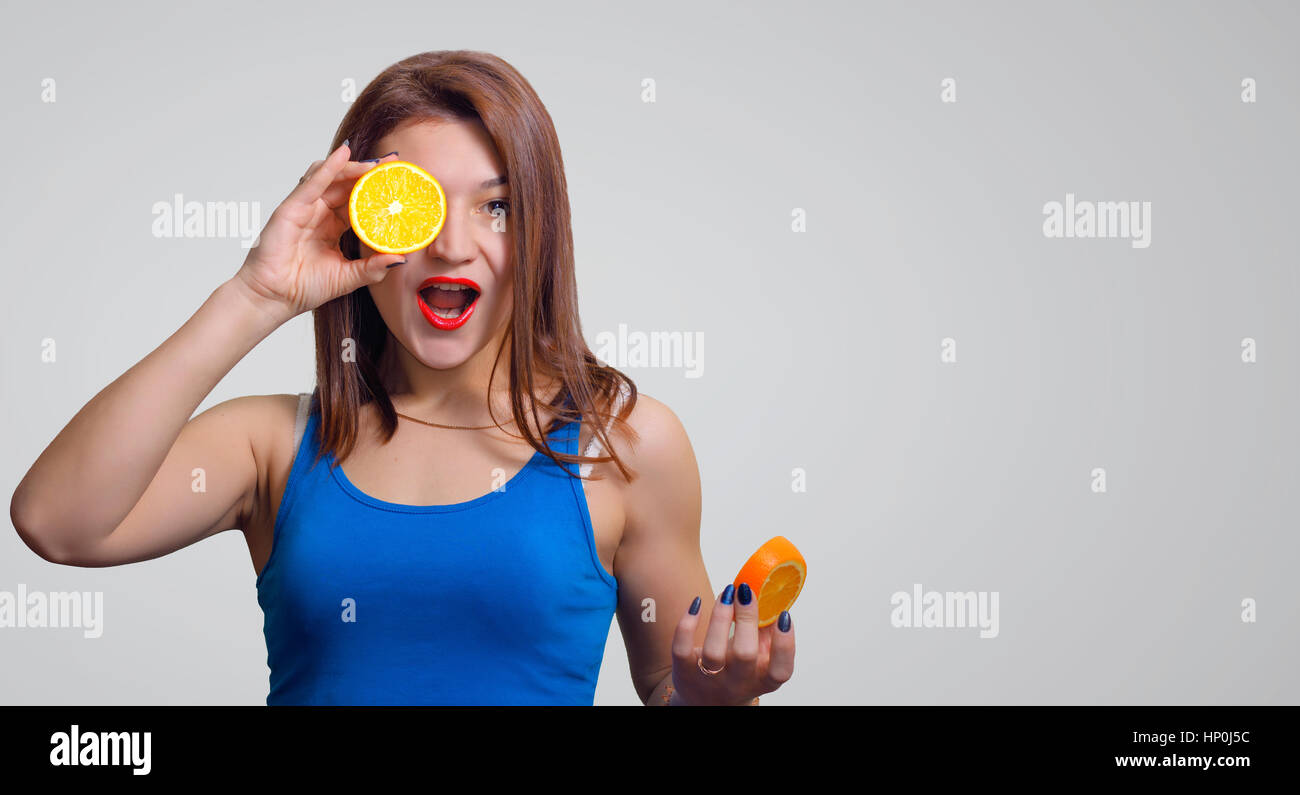 Cheerful young girl avec une bouche ouverte et une tranche d'orange autour des yeux gris sur le fond de studio. Banque D'Images