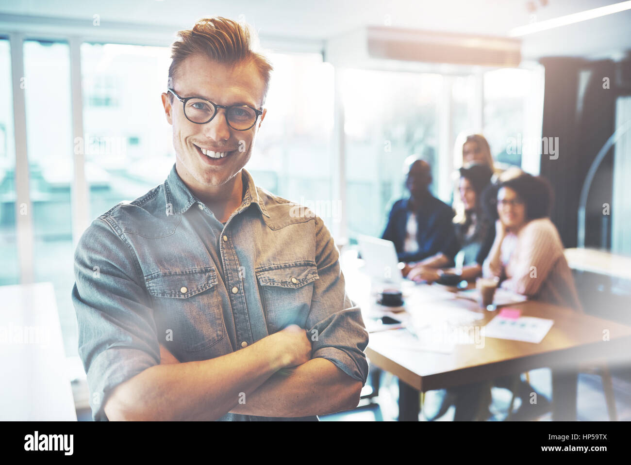 Les jeunes entrepreneurs de confiance dans les verres et les tenues de standing arms folded and smiling in bright office, looking at camera. Son équipe est floue Banque D'Images