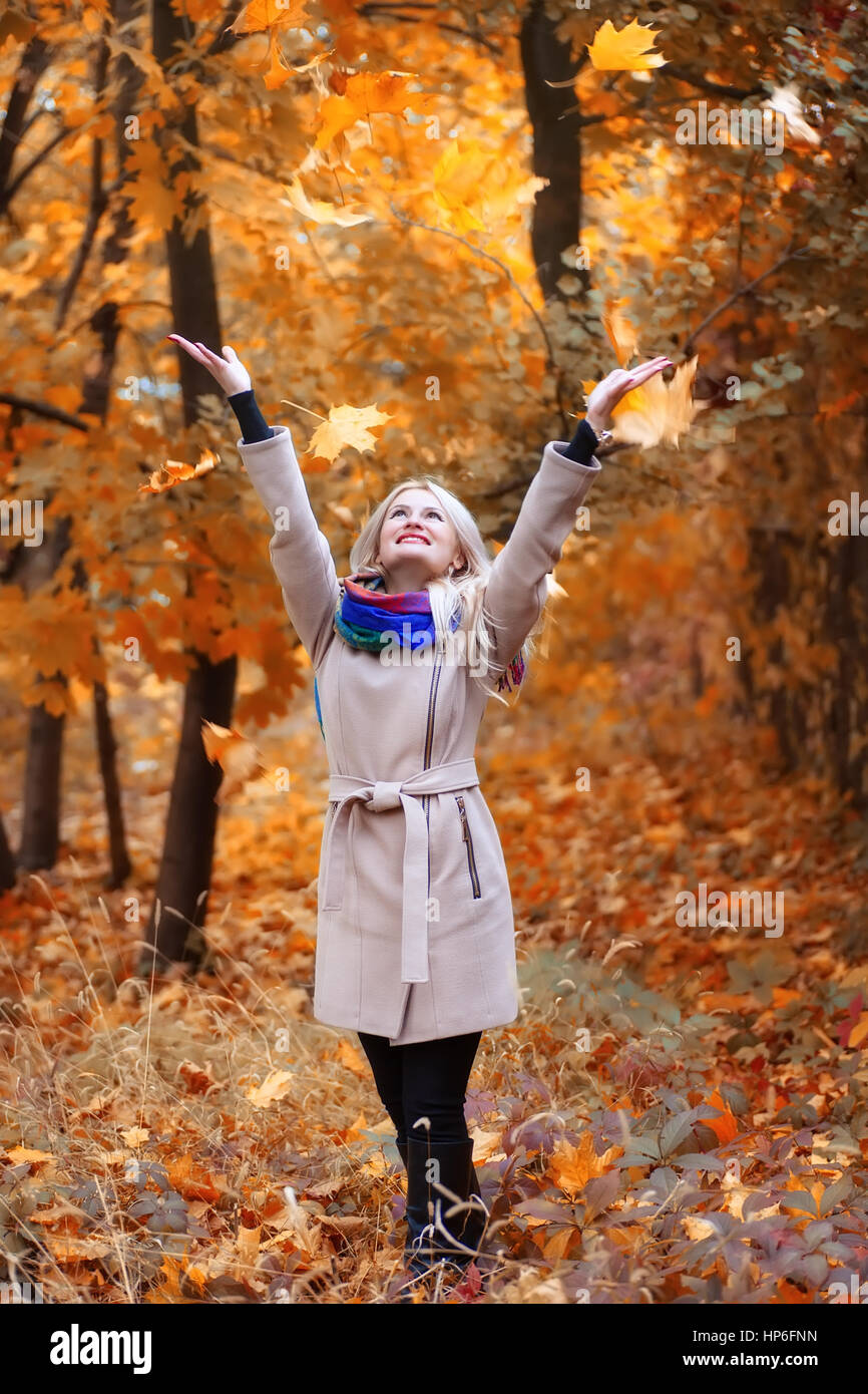 Laughing girl avec de longs cheveux blonds jette feuilles à l'automne parc. Femme heureuse de jouer avec les feuilles d'automne en forêt. Concept loisirs insouciant bonheur Banque D'Images