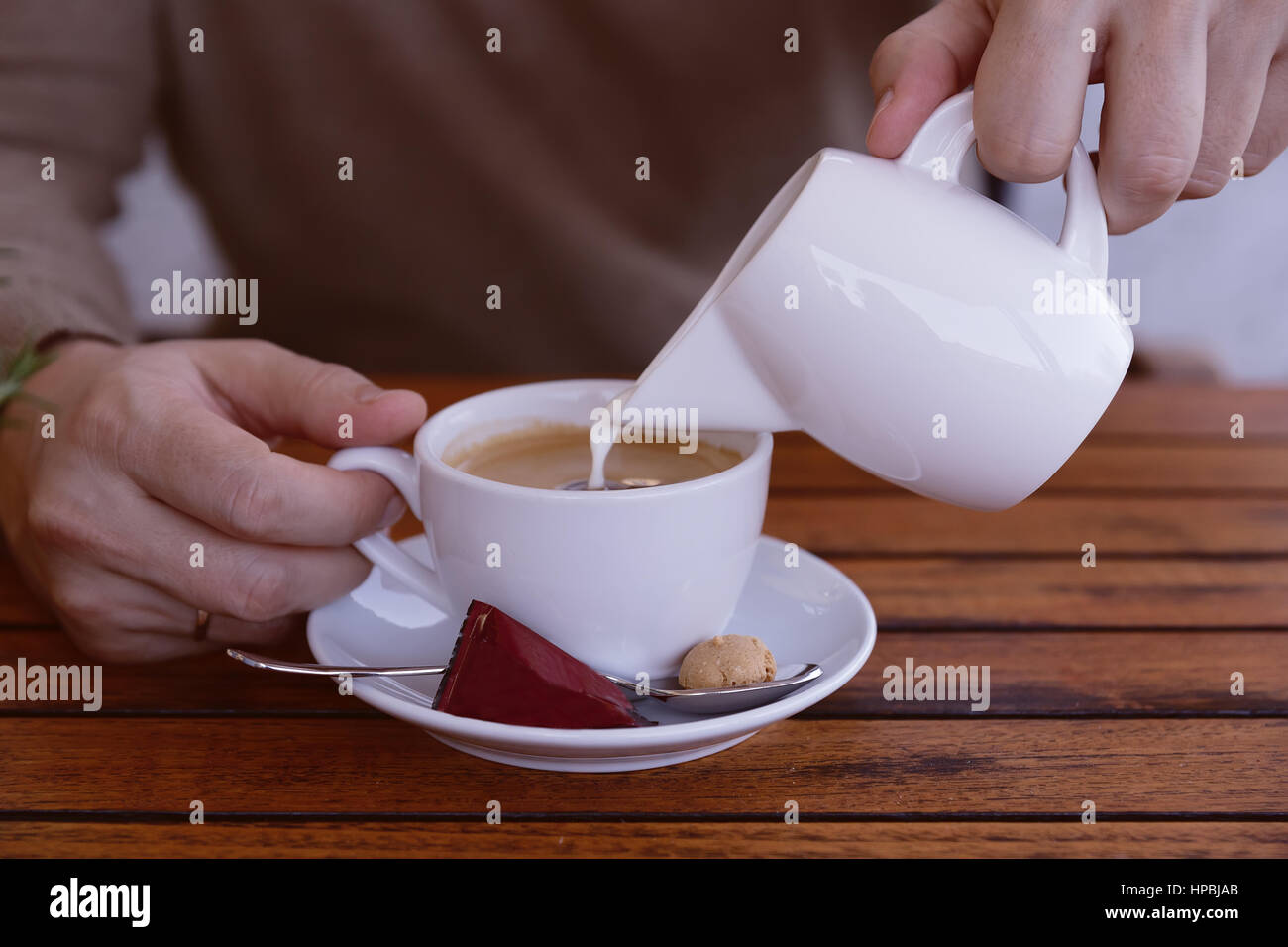 Businessman in pulover verser le lait et la crème à la tasse de café au café-bar ou restaurant sur la table en bois. close-up.Man pouring crème en coupe blanc Banque D'Images