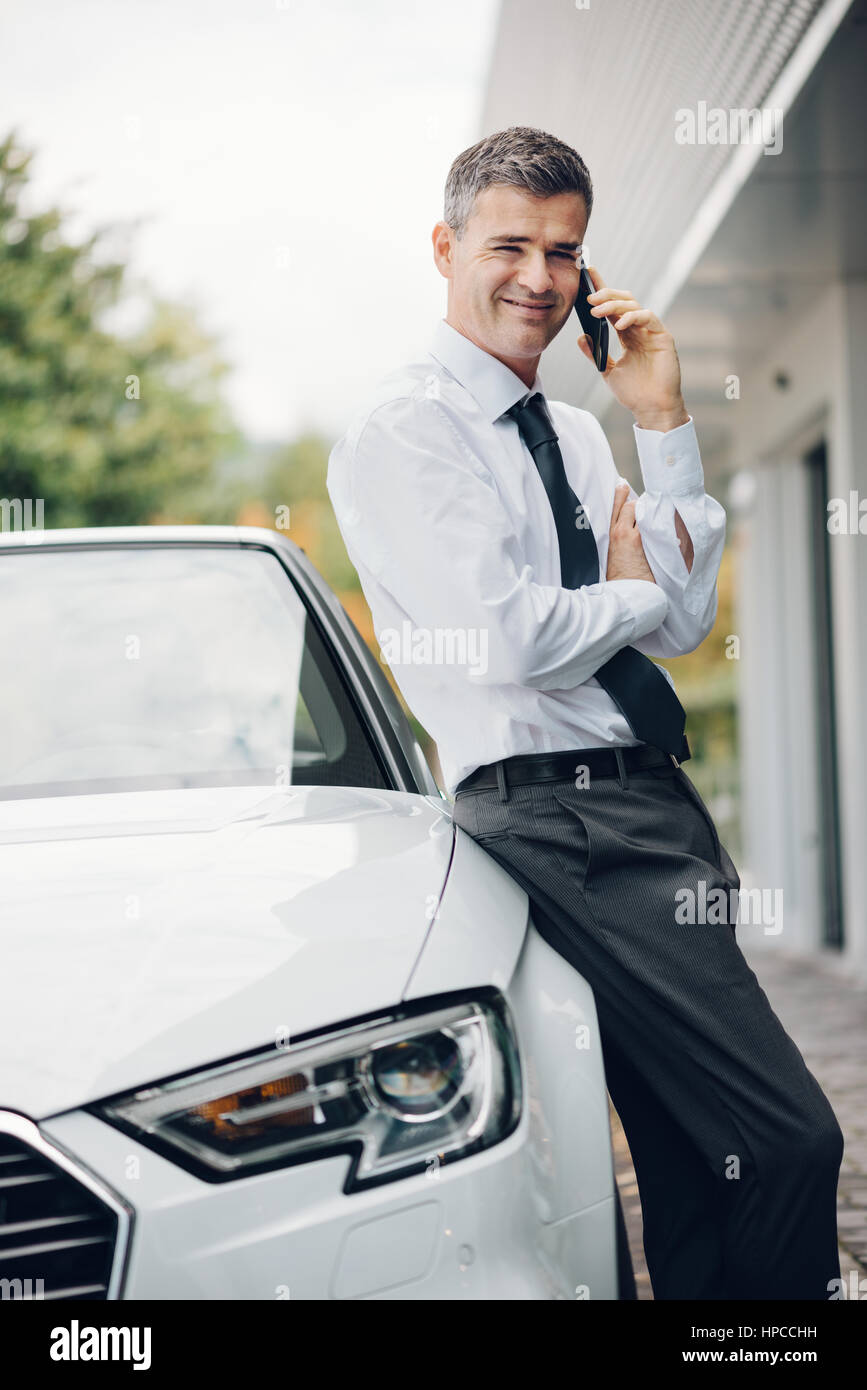 Confiant smiling manager ayant un appel professionnel avec son smartphone et s'appuyant sur son cher location en plein air Banque D'Images