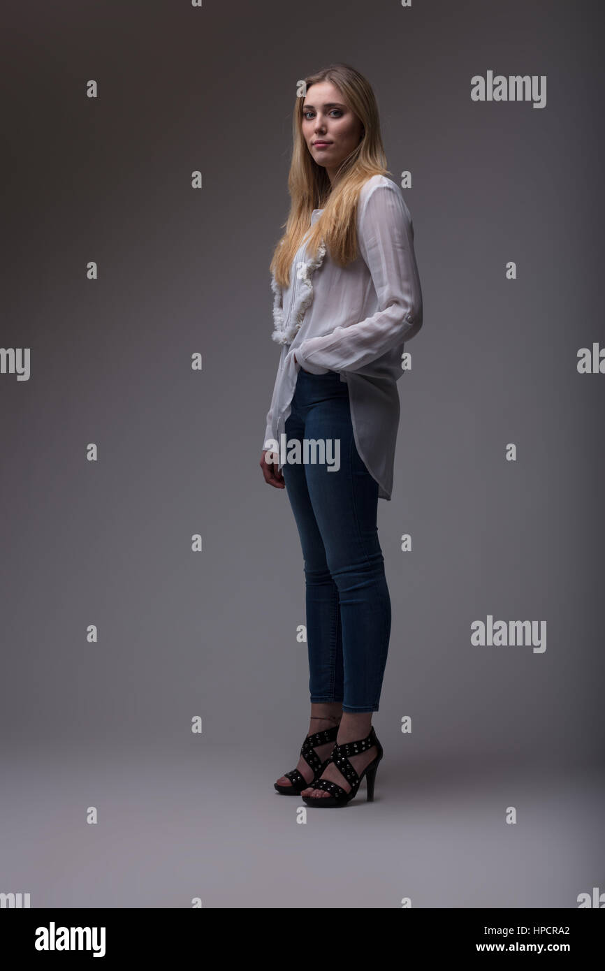 Femme full shot studio portrait, jeune femme avec des jeans bleu denim, ou avec une chemise blanche et de longs cheveux blonds, sur un fond gris d'ombrage Banque D'Images