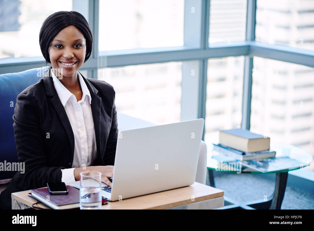 African business woman smiling at camera pendant qu'elle était occupé à travailler sur son ordinateur portable dans le salon affaires de son bureau de travail co moderne, avec de grandes w Banque D'Images