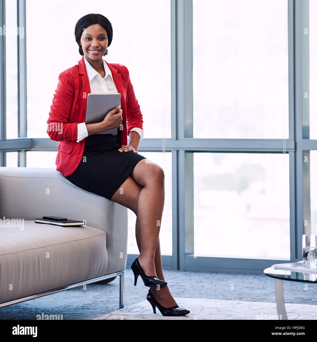 Image de carrés bien habillé noir jeune femme d'entreprise portant un veston  rouge, tenant une tablette numérique tout en étant assis sur le bord de la  table dans un mo Photo Stock -